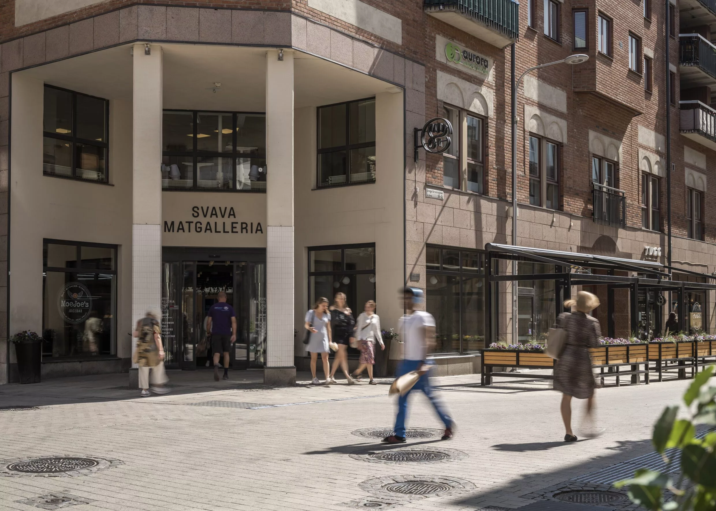 En byggnad med ingången till "Piva Matgalleria" och människor som promenerar utanför på en solig dag.