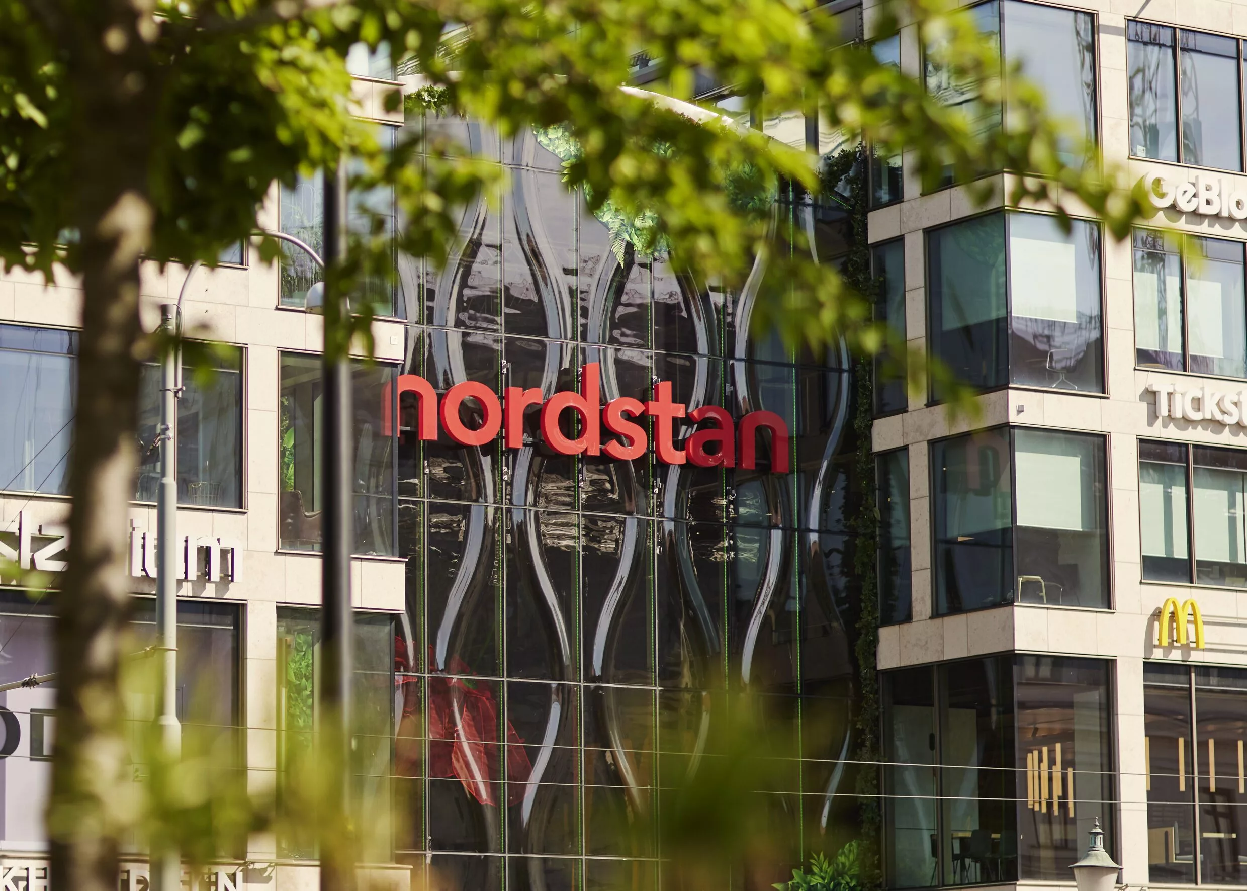 Fasad av Nordstan köpcentrum med glasväggar och röd text "nordstan", omgiven av träd och grönska.