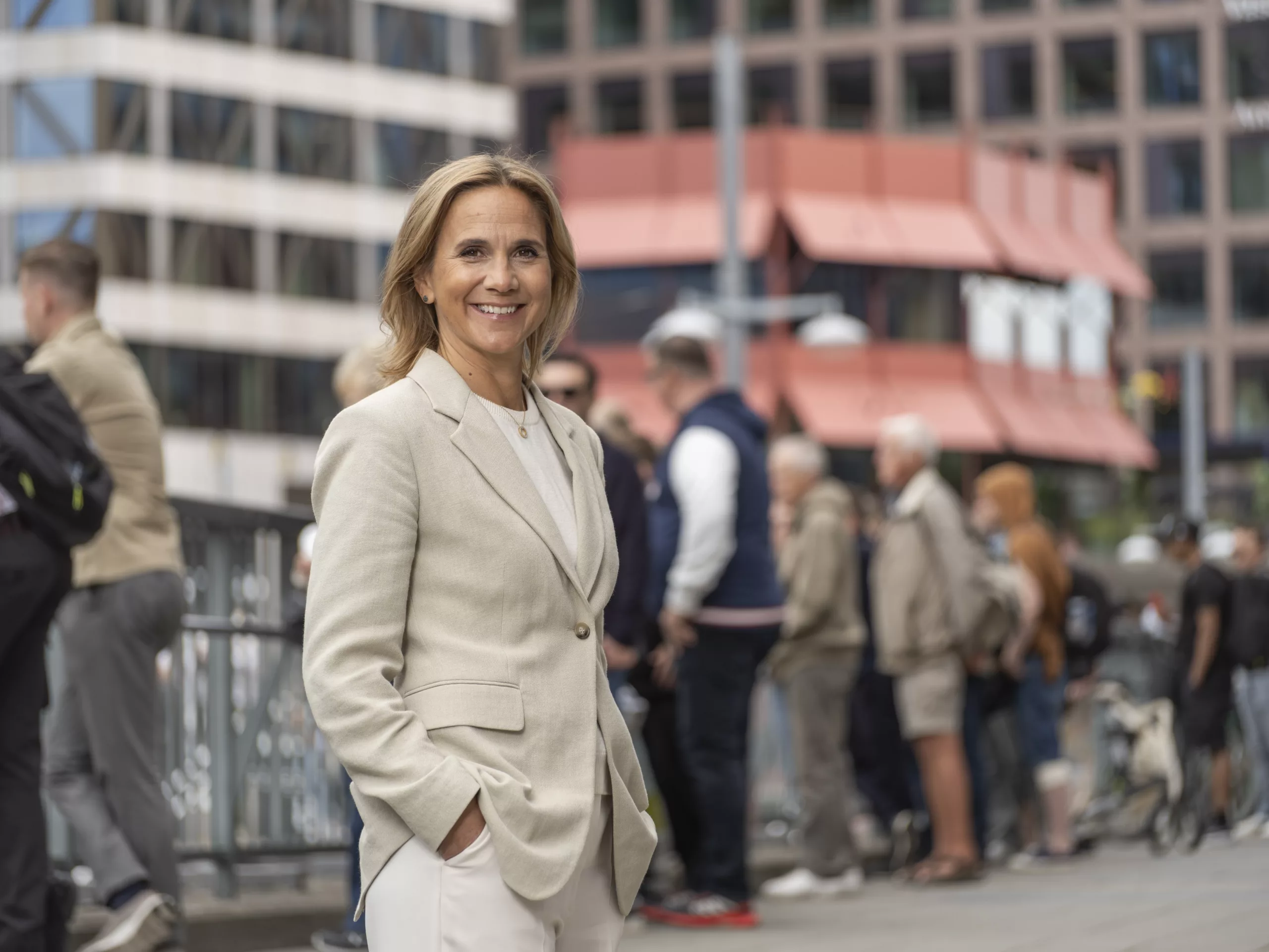 En kvinna i ljus kostym står utomhus med händerna i fickorna; människor och byggnader syns i bakgrunden.
