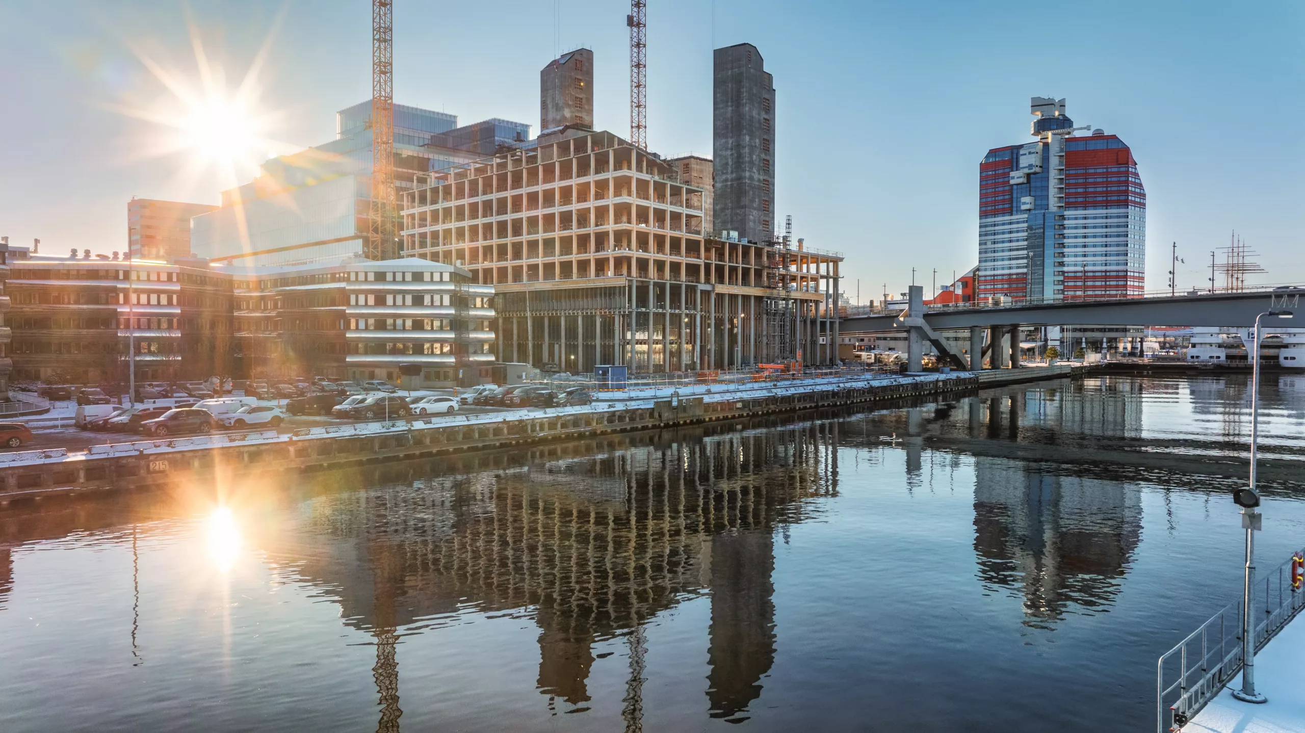 1. Byggarbetsplats vid vattnet med höga byggnader och kranar, reflekterande i vattnet under en solig dag.