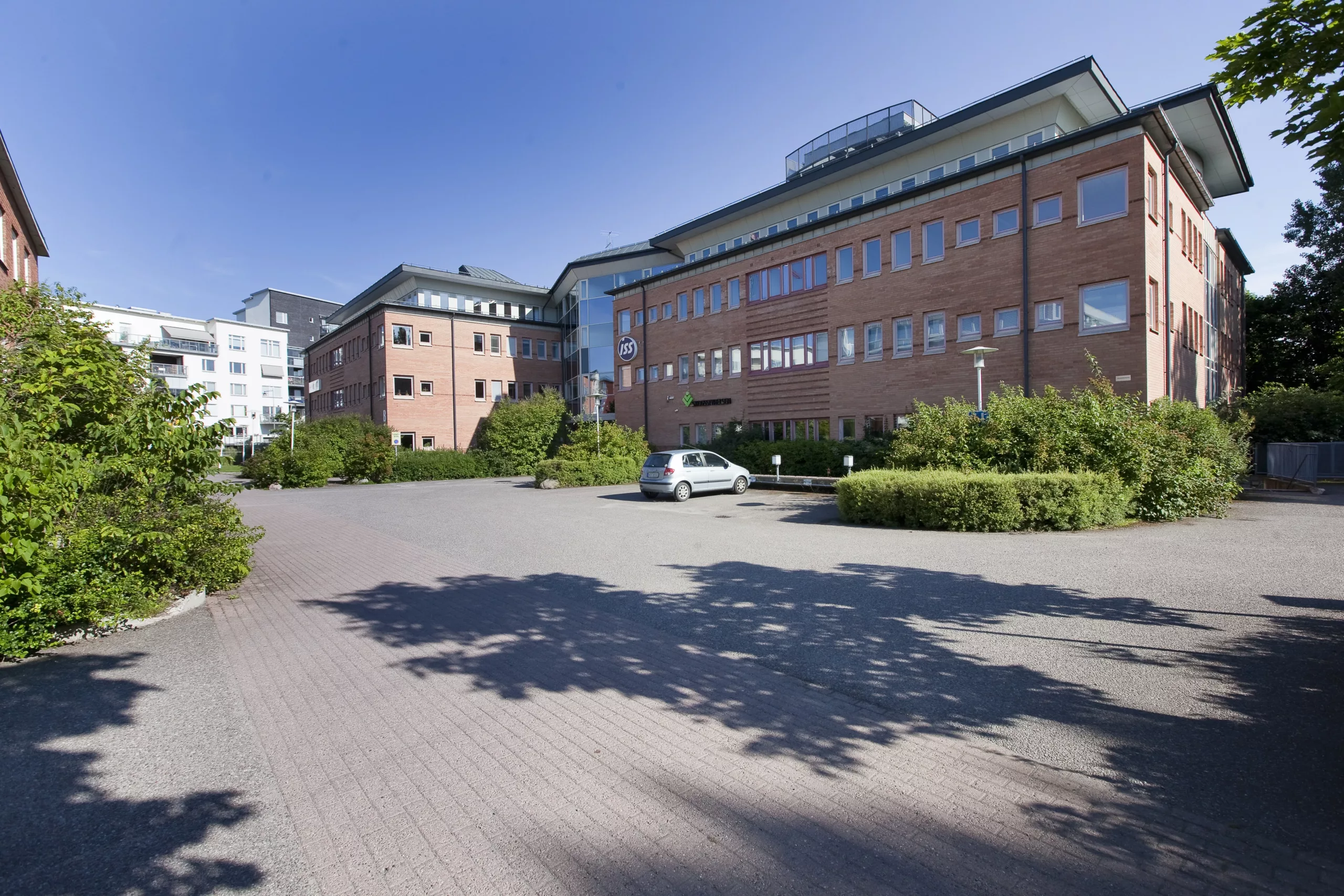 En tegelbyggnad med flera våningar omgiven av buskar och parkeringsplats, under en klar blå himmel.