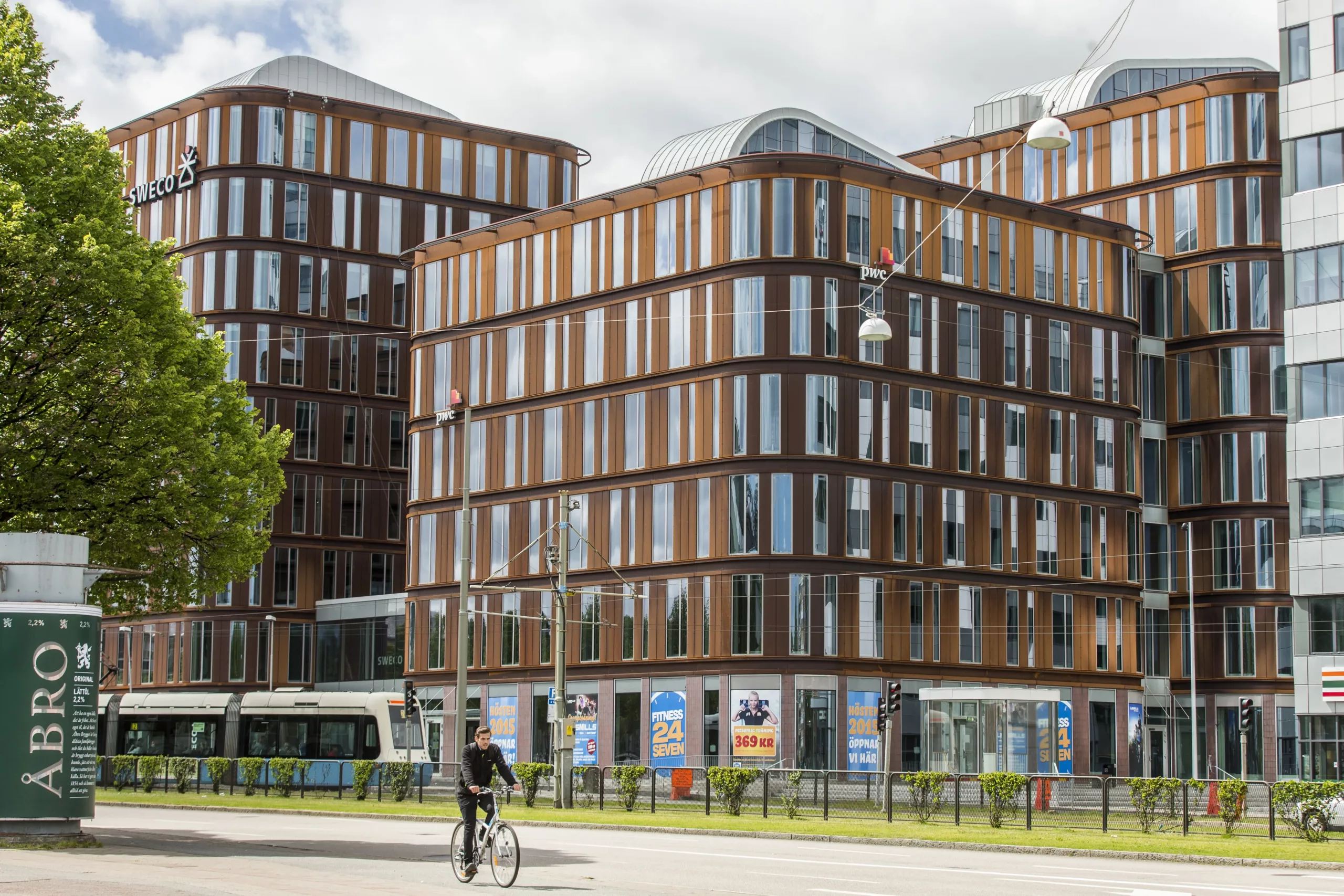 En modern byggnad med fasad av träpaneler och glas, omgiven av en grön trottoar, cyklist och spårvagn i bakgrunden.