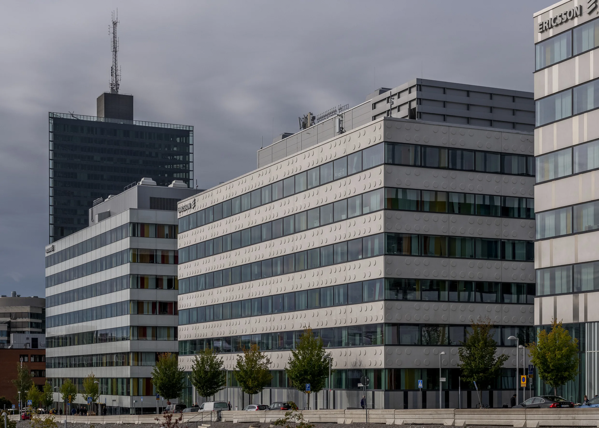 En modern kontorsbyggnad med flera våningar, stora fönster och en höghusbakgrund under en molnig himmel.