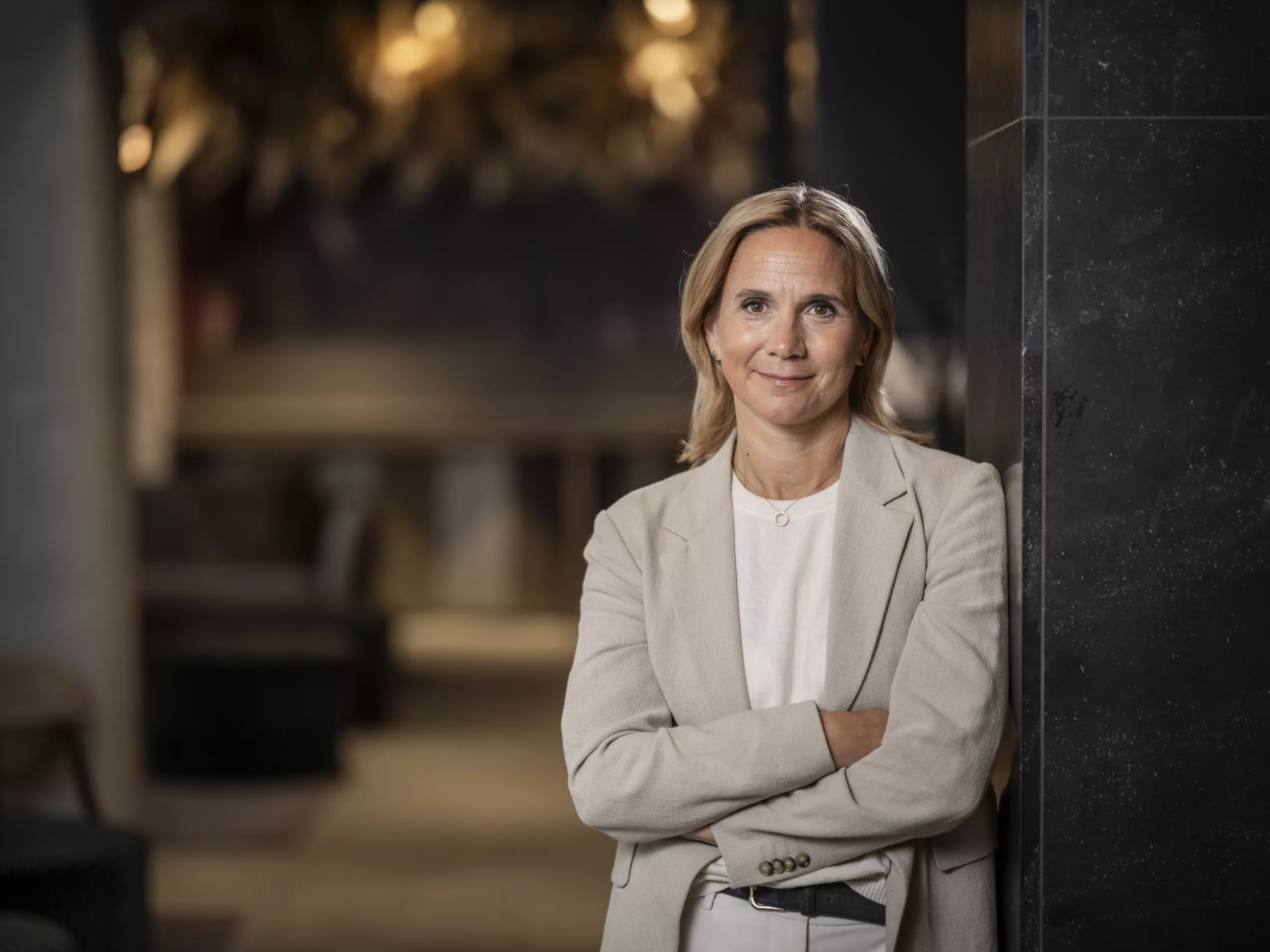 En kvinna i ljus kavaj står med korslagda armar i en modern inomhusmiljö med dämpad belysning.
