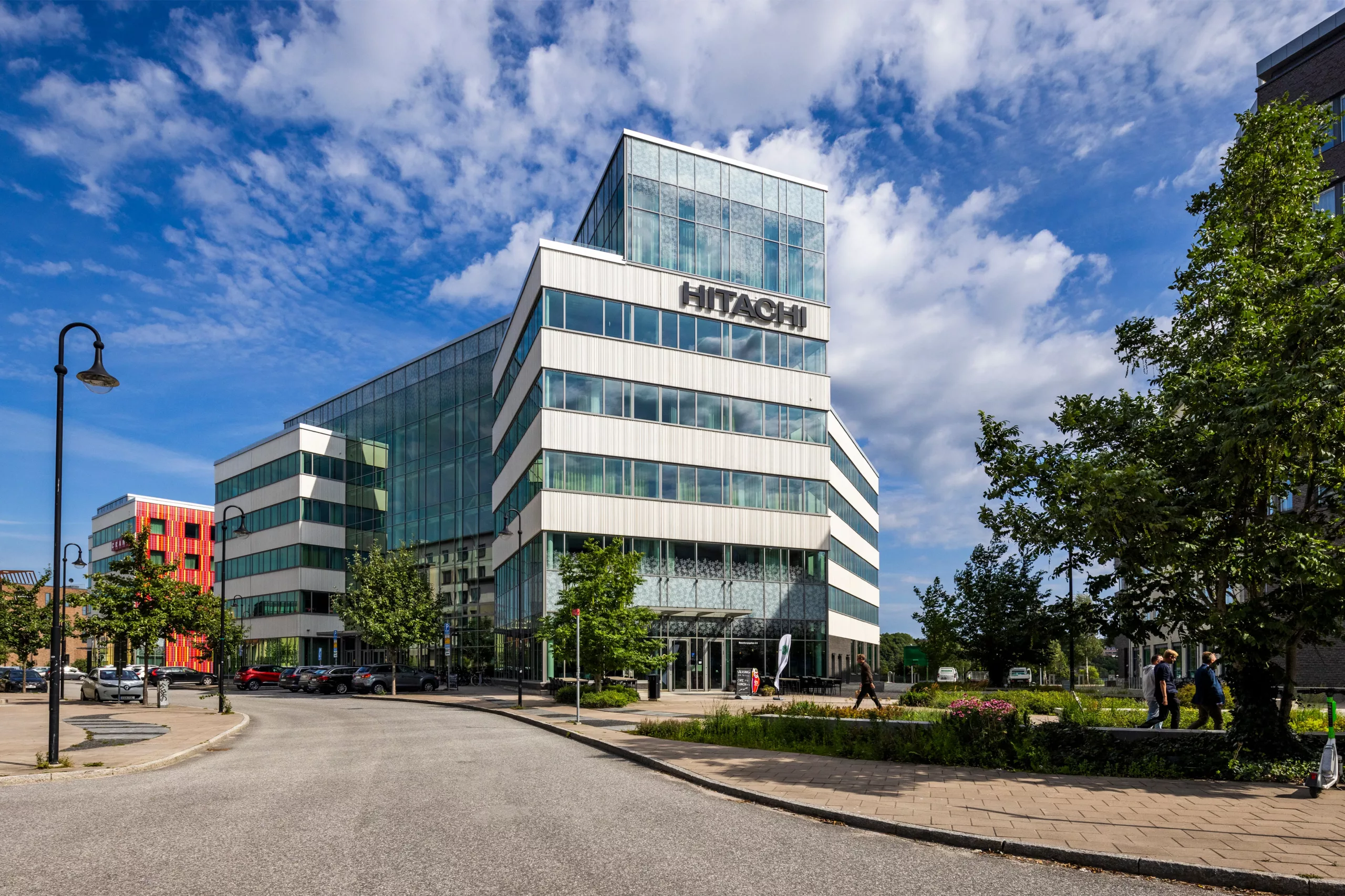 En glasfasad byggnad med texten "Hitachi" på toppen, omgiven av grönska och en klar himmel.