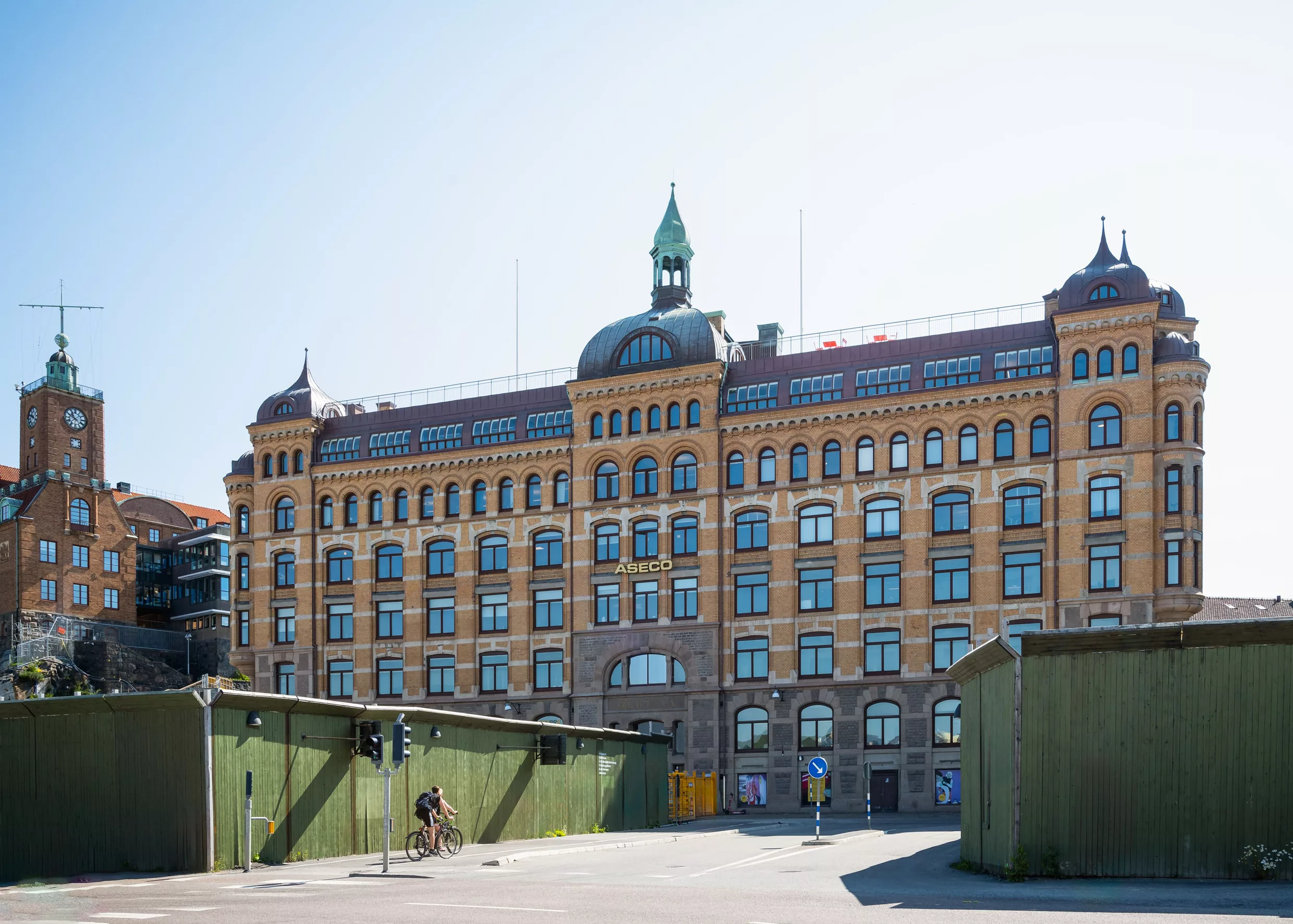 En stor gul byggnad med flera våningar och grönt torn på taket, omgiven av byggnadsställningar och grön plank.