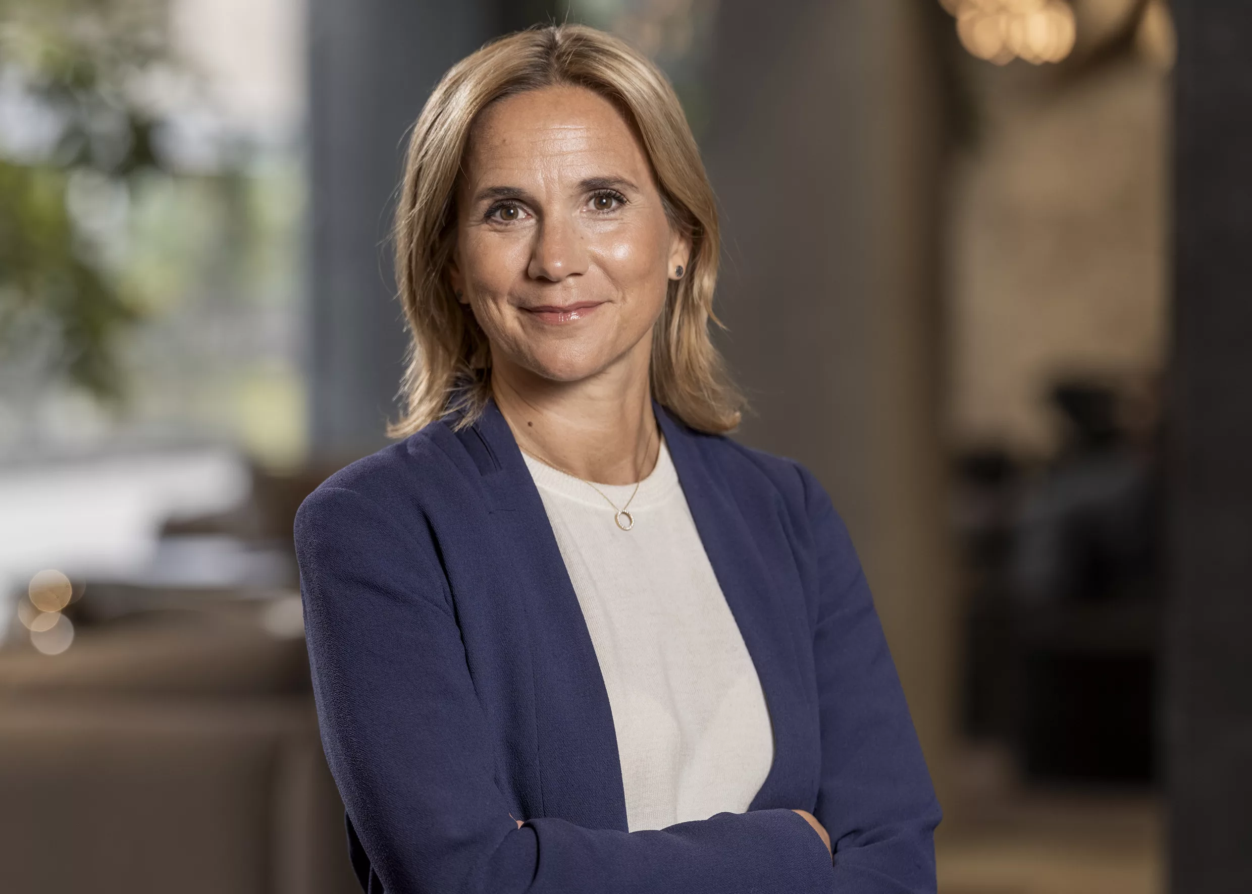 En kvinna i blå kavaj och vit topp som står med korslagda armar och ler, med en suddig inomhusmiljö i bakgrunden.