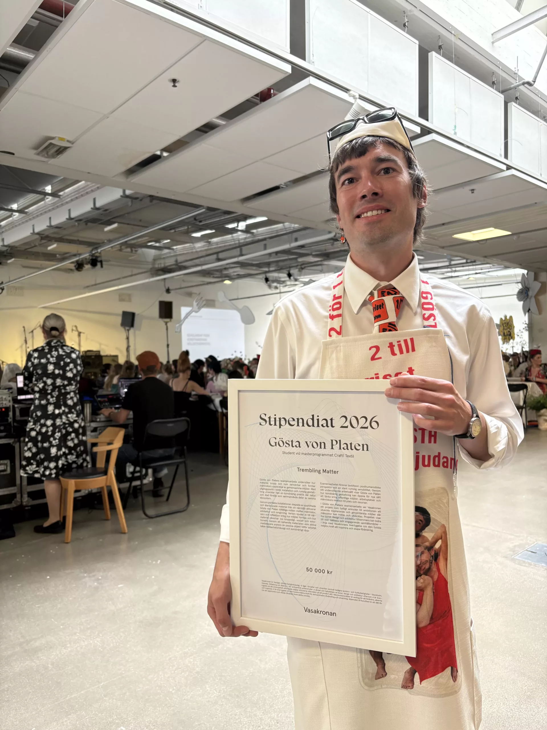 En person i en festlig outfit håller upp ett diplom med texten "Skolmästare 2023" framför en matsal med gäster.