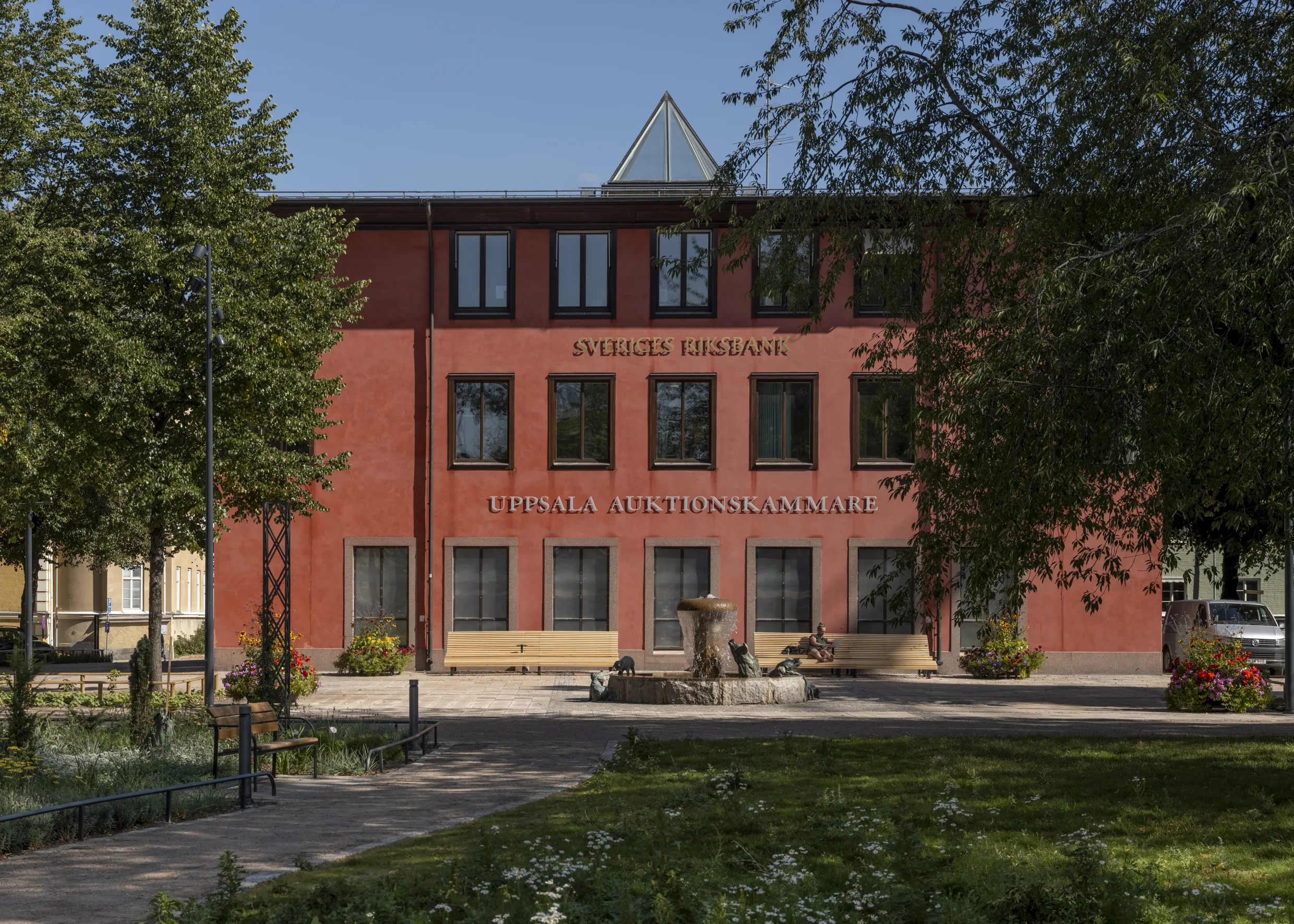 En röd byggnad med texten "Östra Real Gymnasium" synlig, omgiven av träd och en grön park.