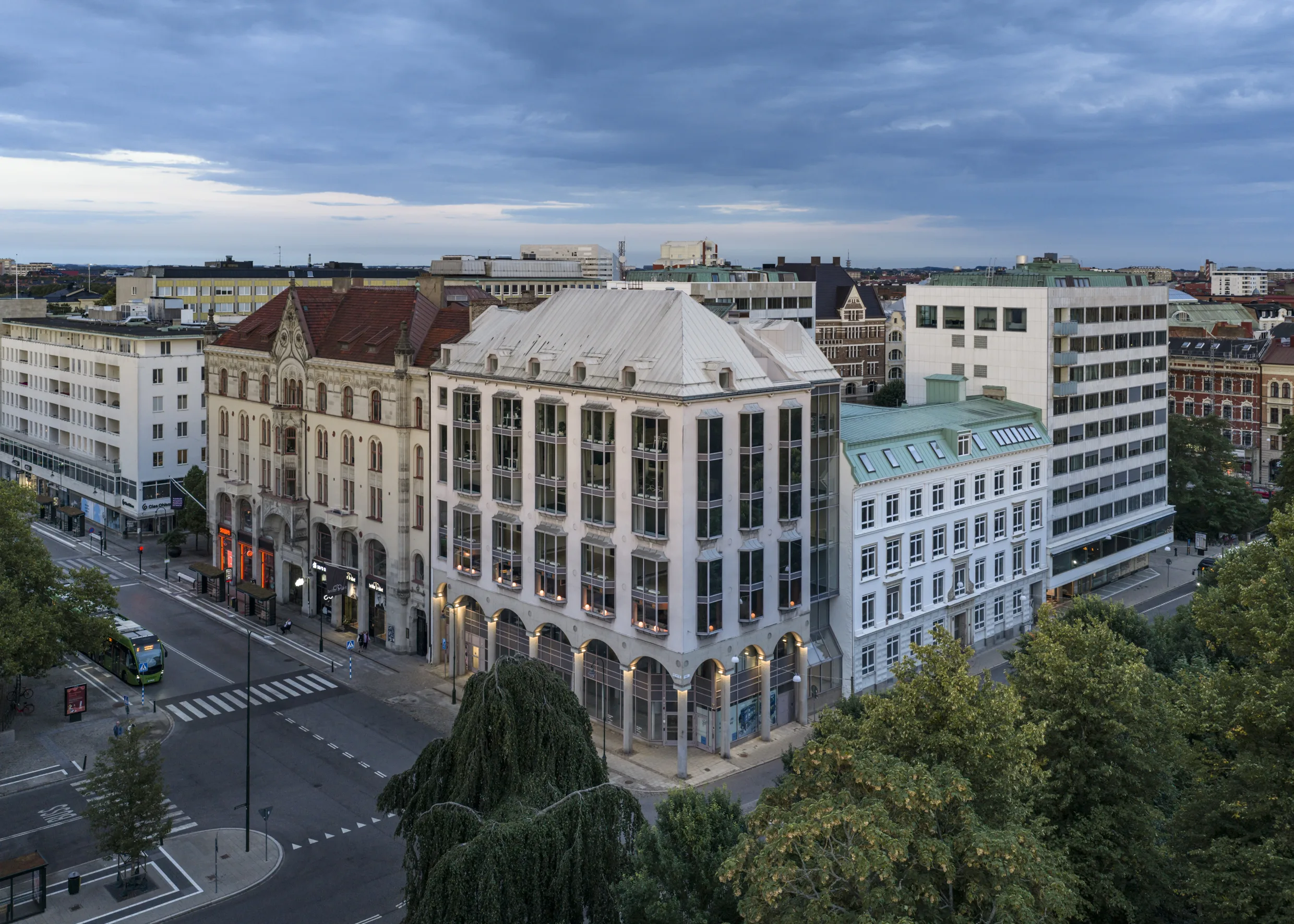 Flera byggnader i klassisk arkitektur vid en korsning, omgivna av träd och fotograferade under en molnig himmel.
