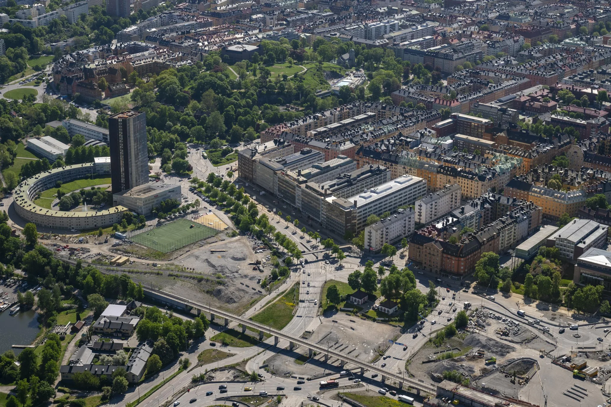 Flygfoto över en stadsdel med byggnader, vägar och grönområde i bakgrunden.