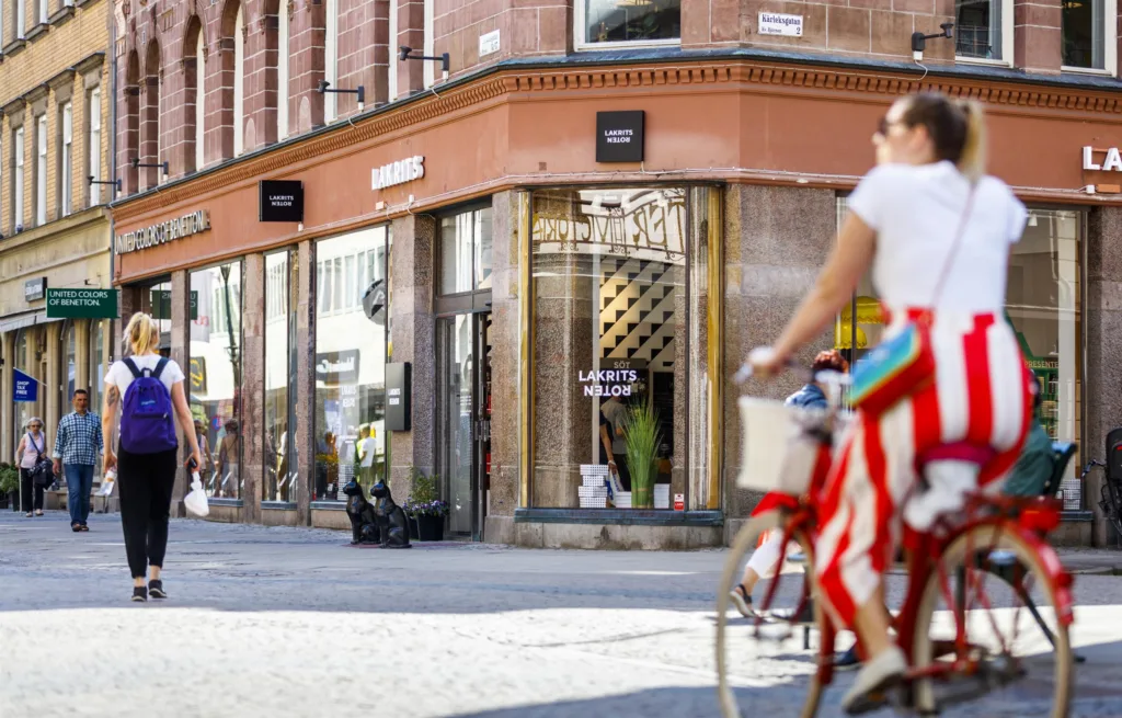 En person i vit topp och randiga byxor cyklar förbi en butik med stora glasfönster vid en trottoar.