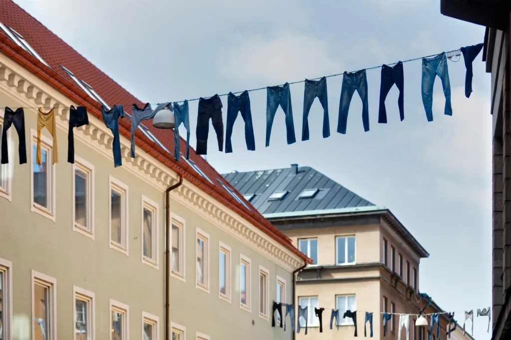 Jeans hänger på klädstreck mellan byggnader, med tak och fönster i bakgrunden.
