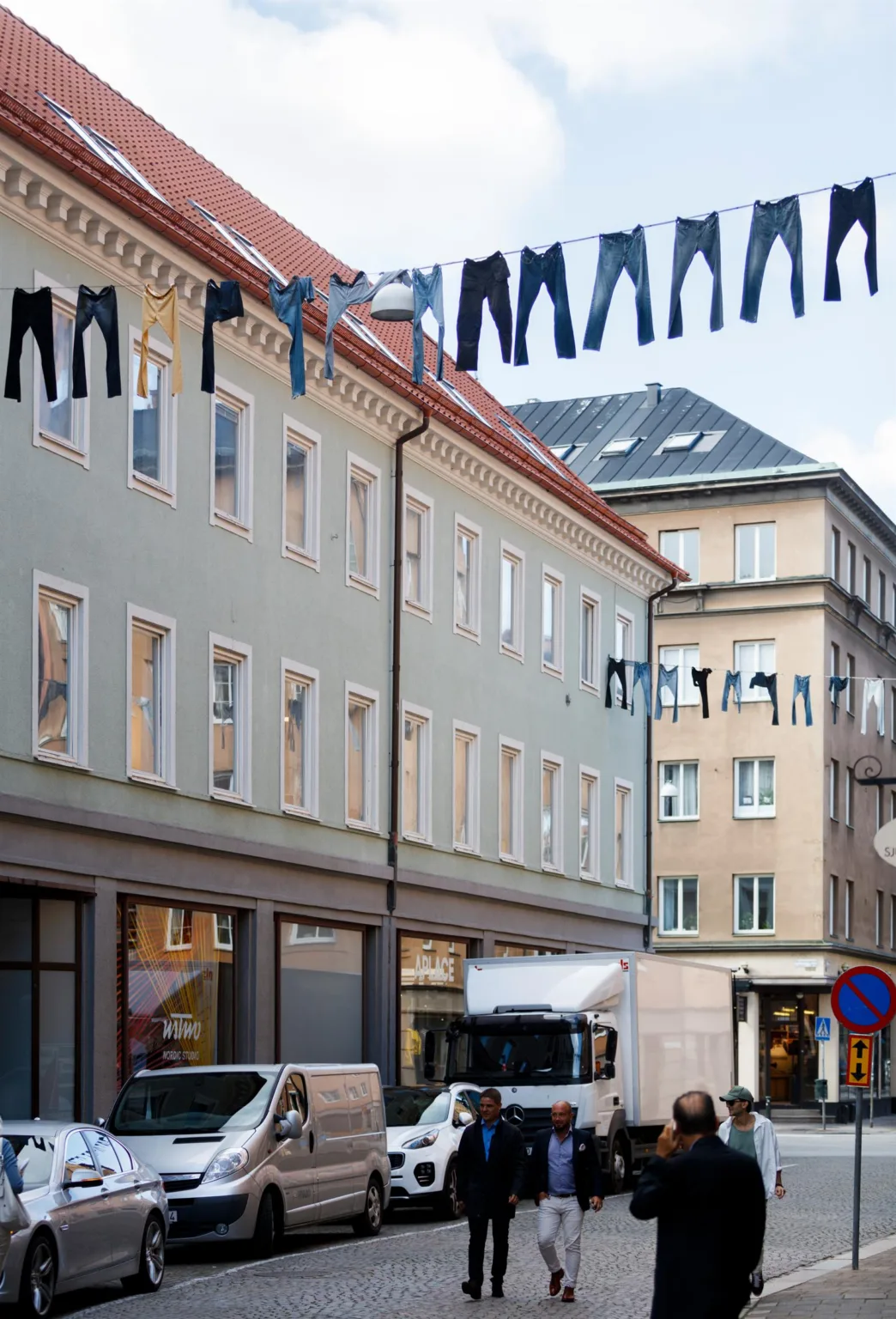 Jeans hänger på klädstreck mellan byggnader med butiksfönster och en parkerad bil nedanför.