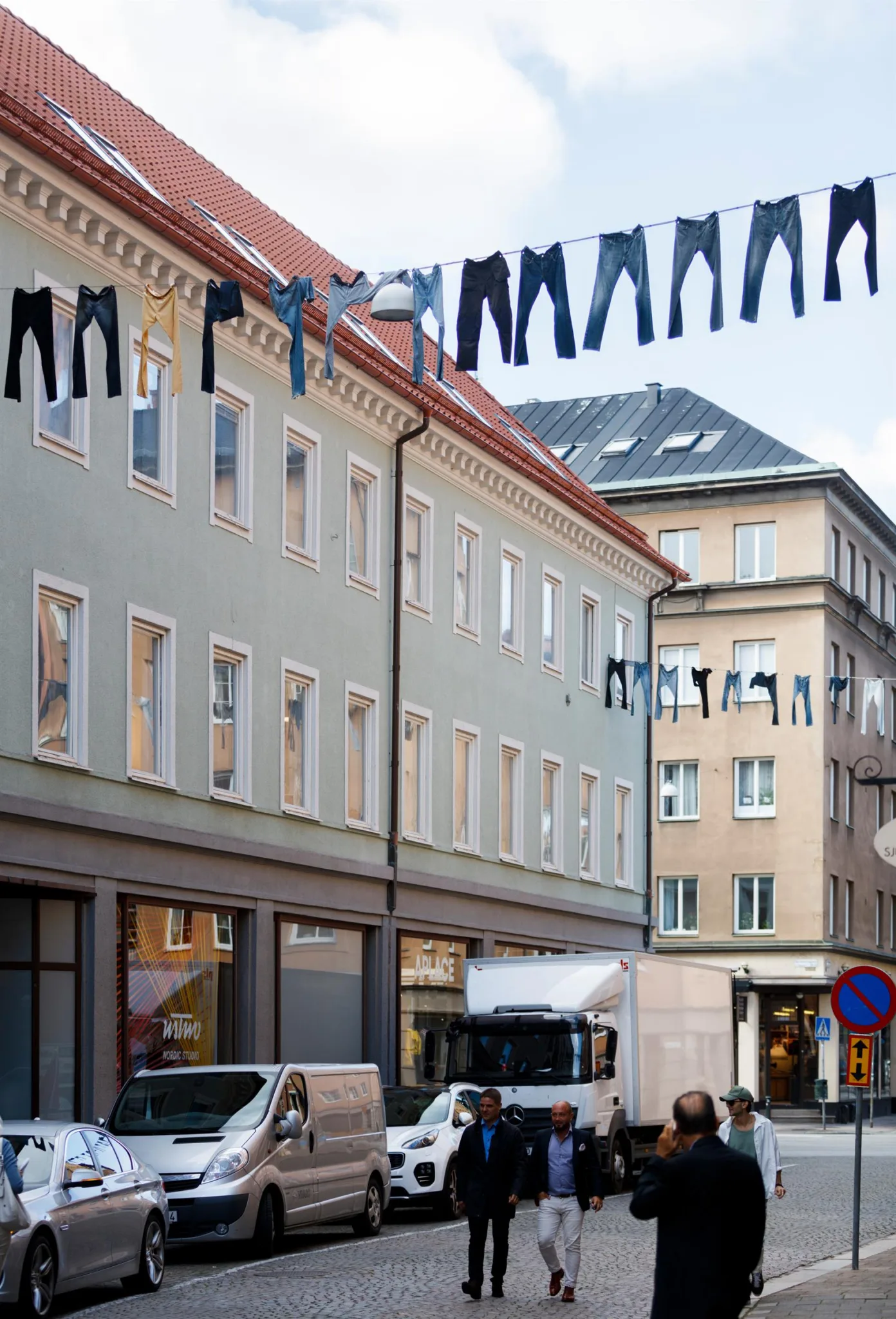 Jeans hänger på klädstreck mellan byggnader med butiksfönster och en parkerad bil nedanför.
