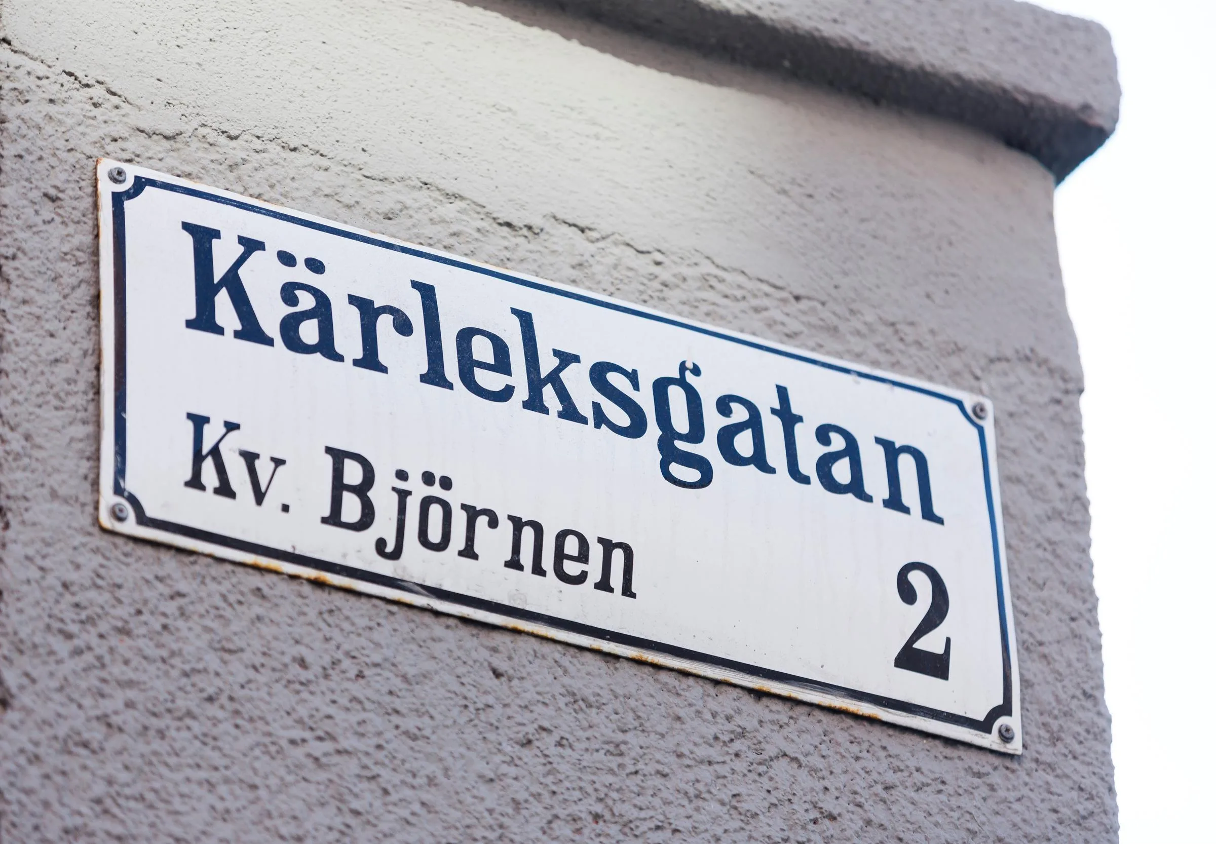 En vit gatunamnskylt på en grå vägg med texten "Kärleksgatan, Kv. Björnen, 2".