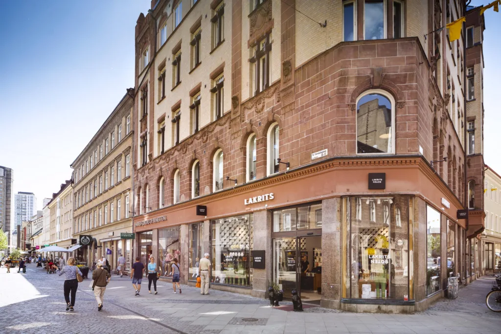 En rödbrun stenbyggnad med stora skyltfönster och människor som går längs trottoaren.