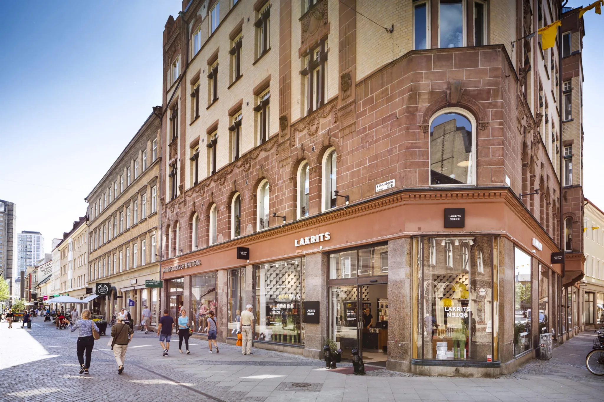 En rödbrun stenbyggnad med stora skyltfönster och människor som går längs trottoaren.