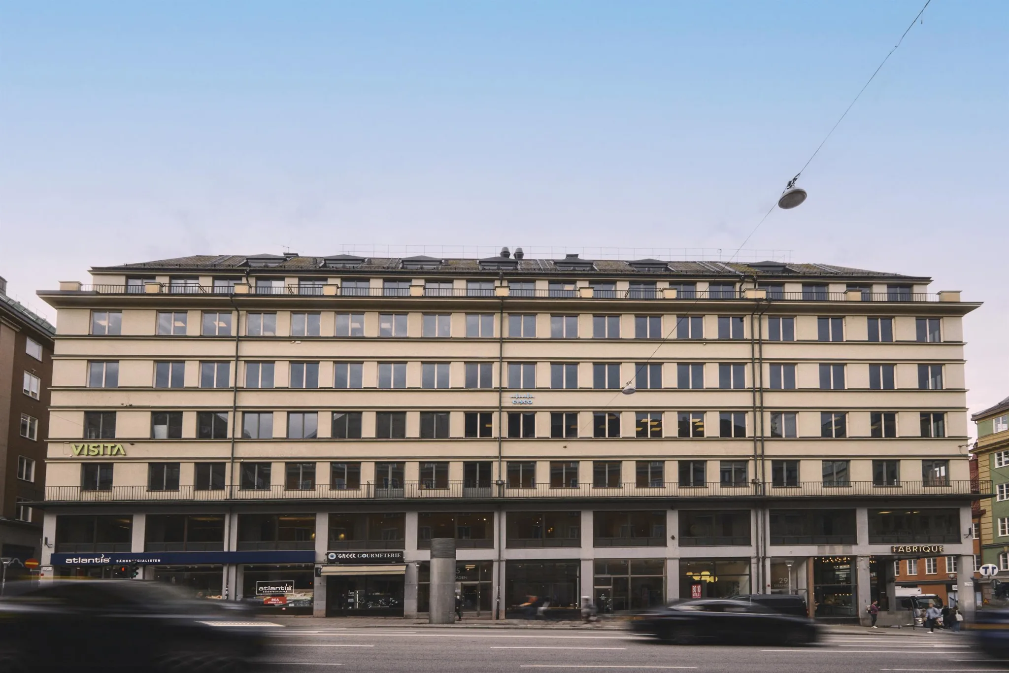 En gul byggnad med flera våningar och stora fönster, omgiven av en bred gata med bilar.