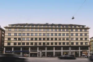 En gul byggnad med flera våningar och stora fönster, omgiven av en bred gata med bilar.
