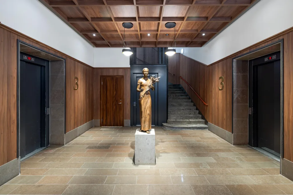 En staty av en person på en piedestal i en elegant entréhall med trappor och träpaneler på väggarna.