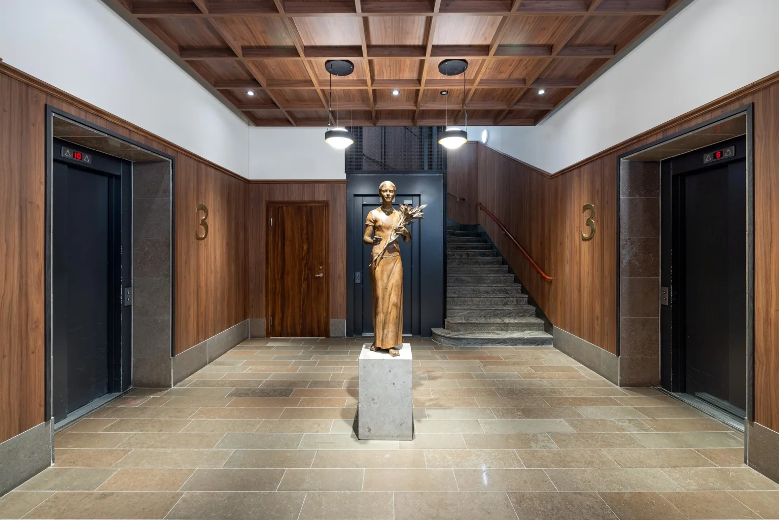 En staty av en person på en piedestal i en elegant entréhall med trappor och träpaneler på väggarna.