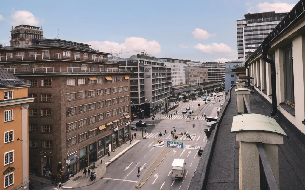 En stadsvy med en bred gata kantad av höga byggnader och fotgängare som rör sig längs trottoaren.