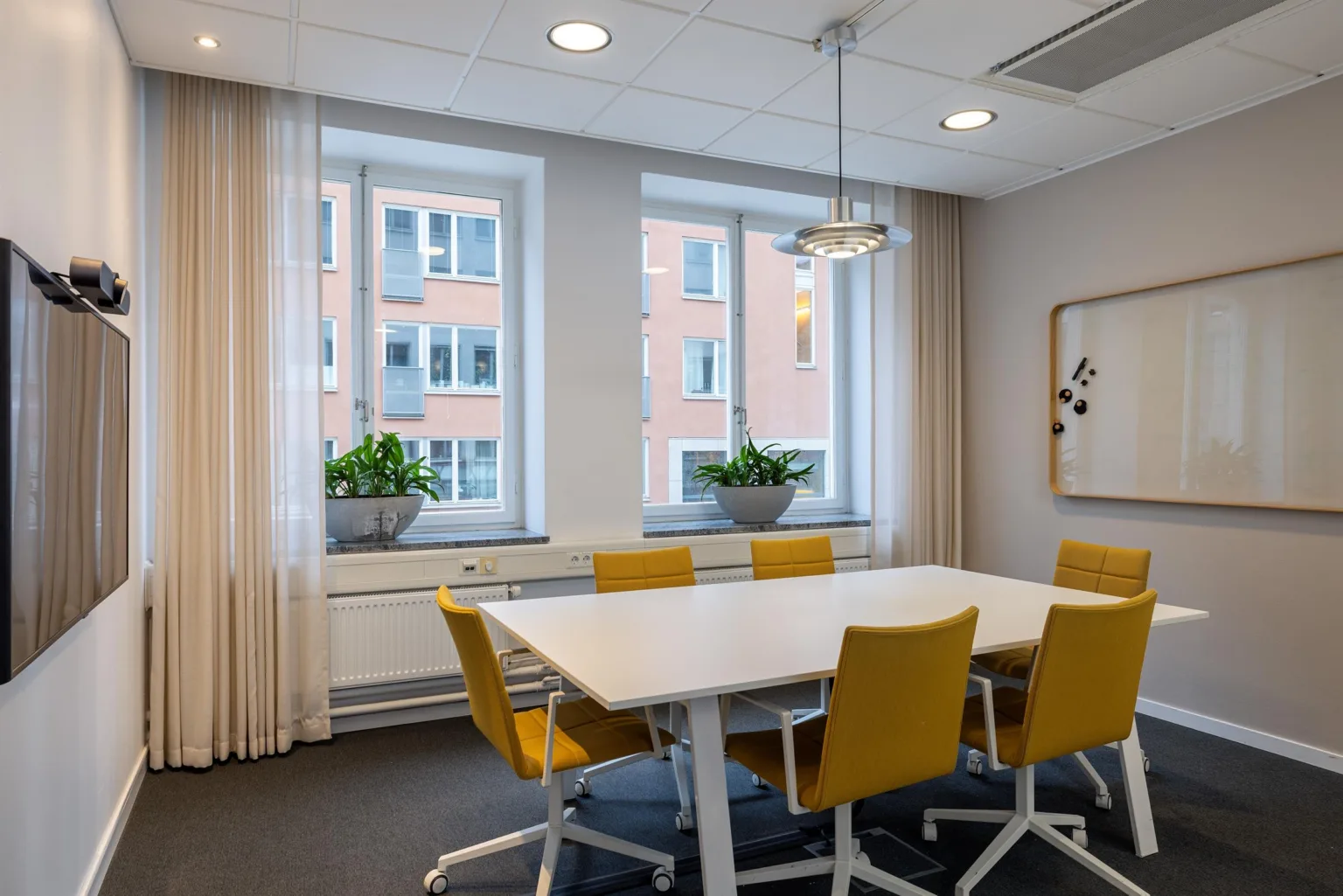 5. Ett mindre mötesrum med ett vitt bord omgivet av gula stolar, två fönster med växter på fönsterbrädorna och en anslagstavla på väggen.