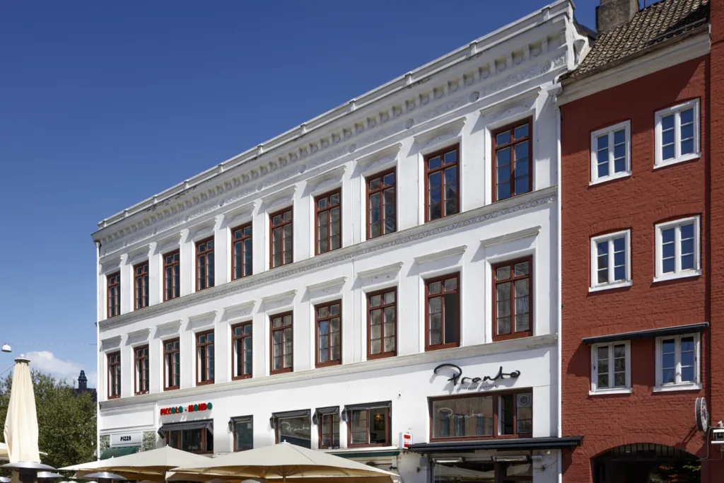 Vit byggnad med stora fönster, en restaurang med uteservering synlig längst ner.