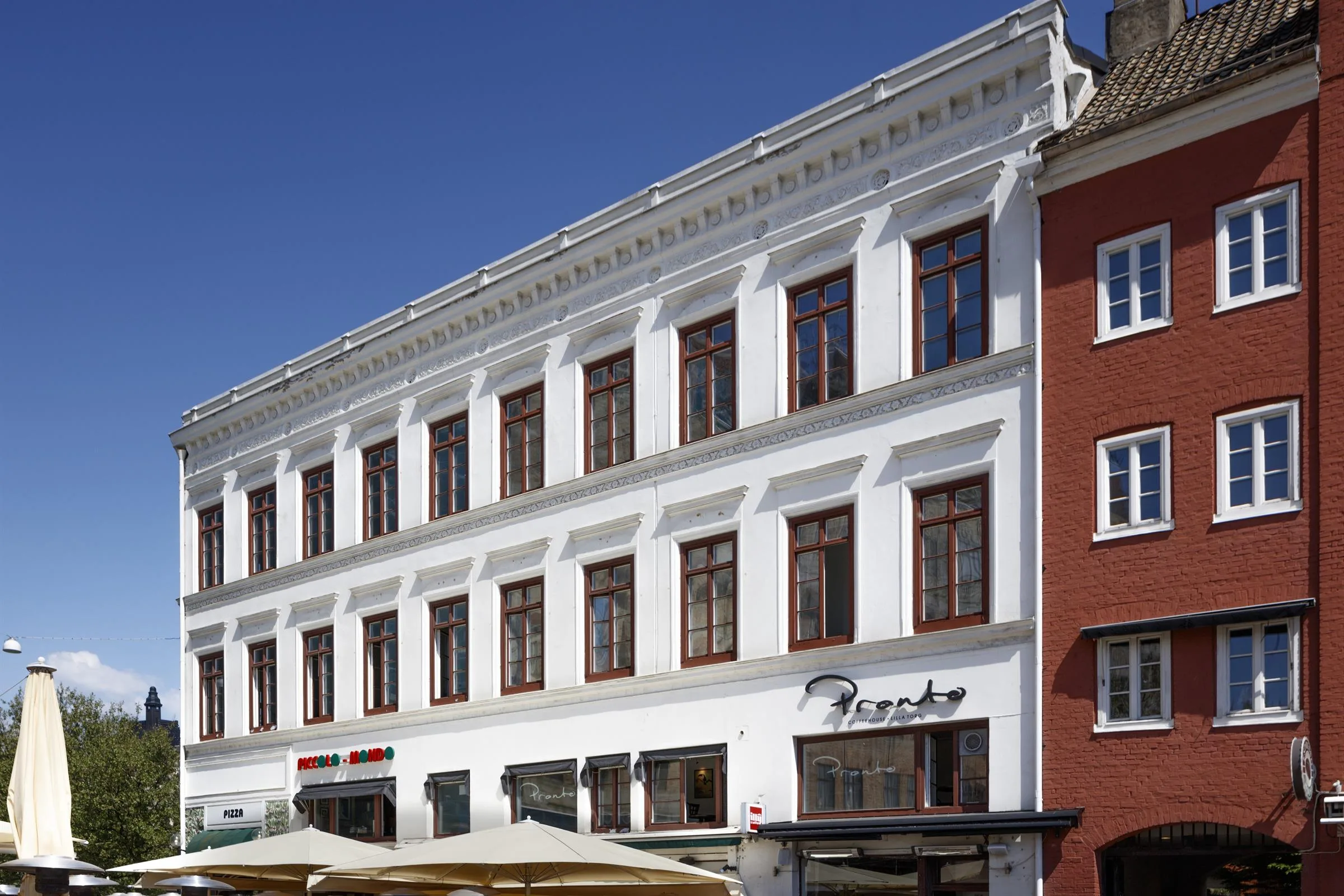 Vit byggnad med stora fönster, en restaurang med uteservering synlig längst ner.