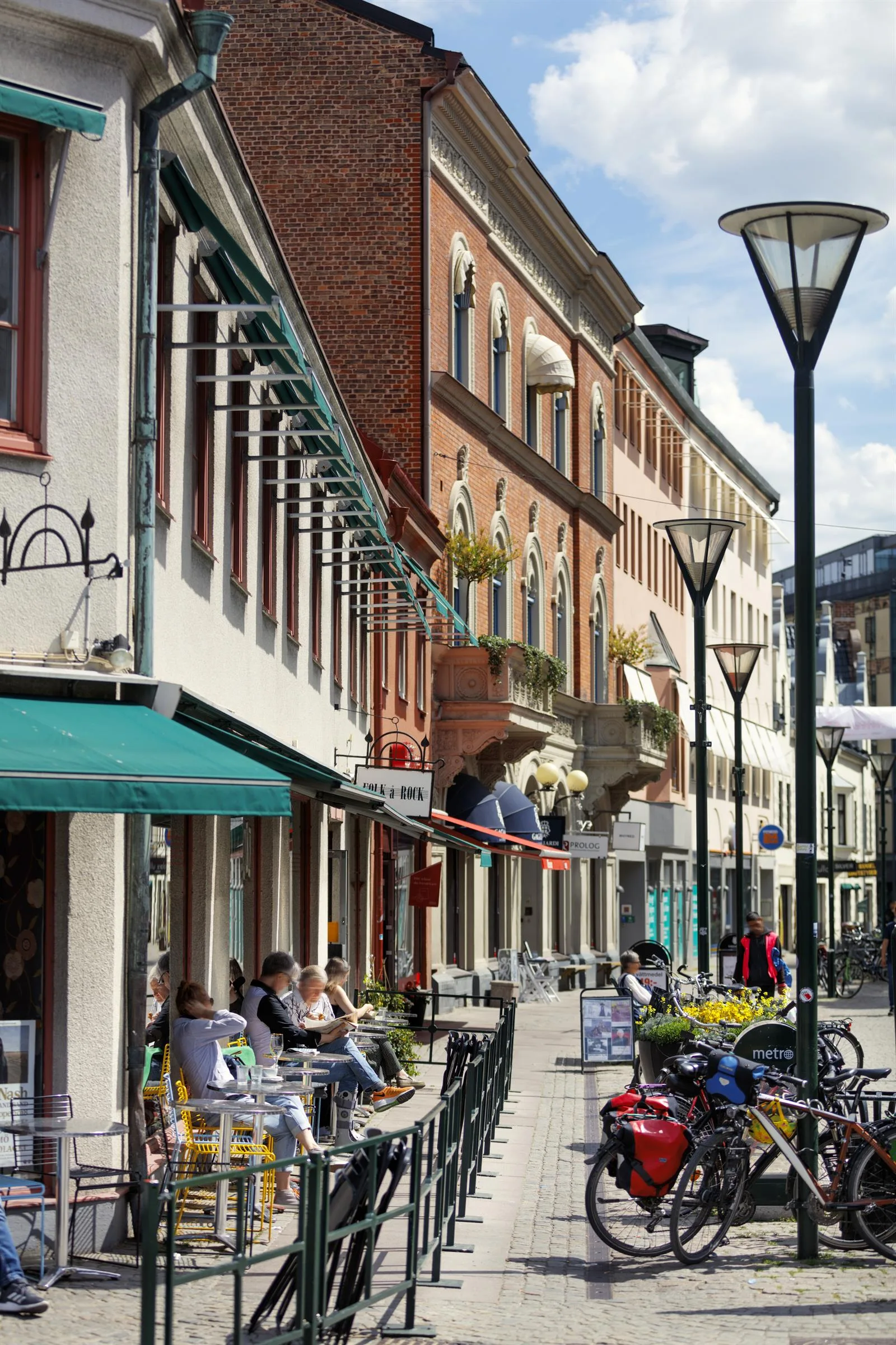 Gågata med uteserveringar, cyklar och människor framför färgglada butiksfasader.