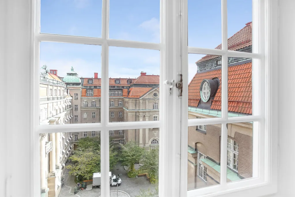 Ett öppet fönster med utsikt över en innergård och tak med röda tegelpannor.