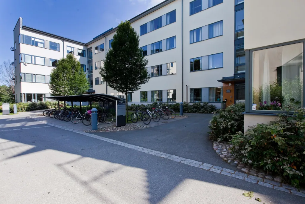 En bostadsbyggnad med cykelställ och grönska framför, omgiven av gångvägar.