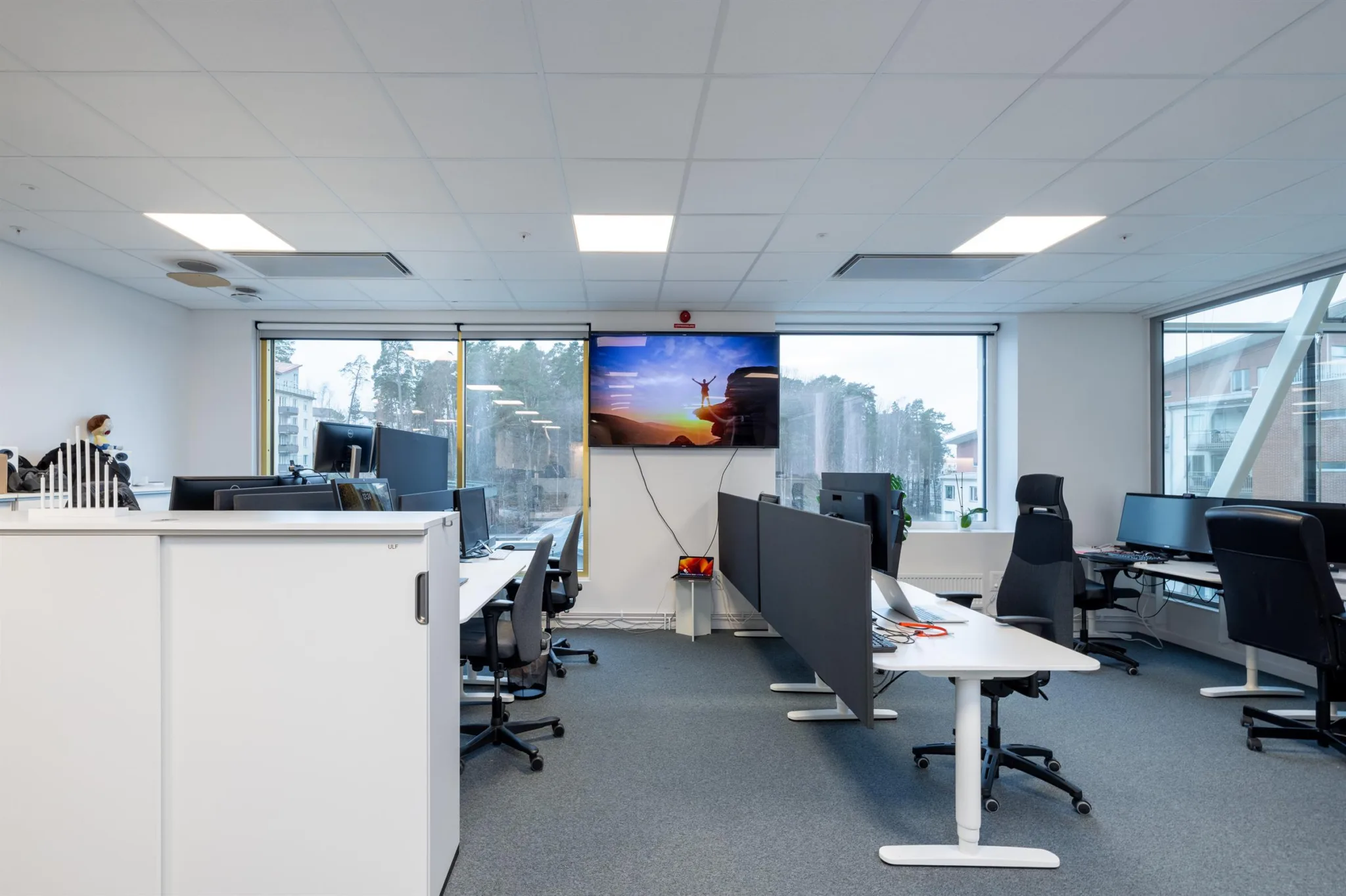 Ett kontorsutrymme med skrivbord, skärmar och kontorsstolar, samt en stor väggmonterad TV som visar en färgglad bild.