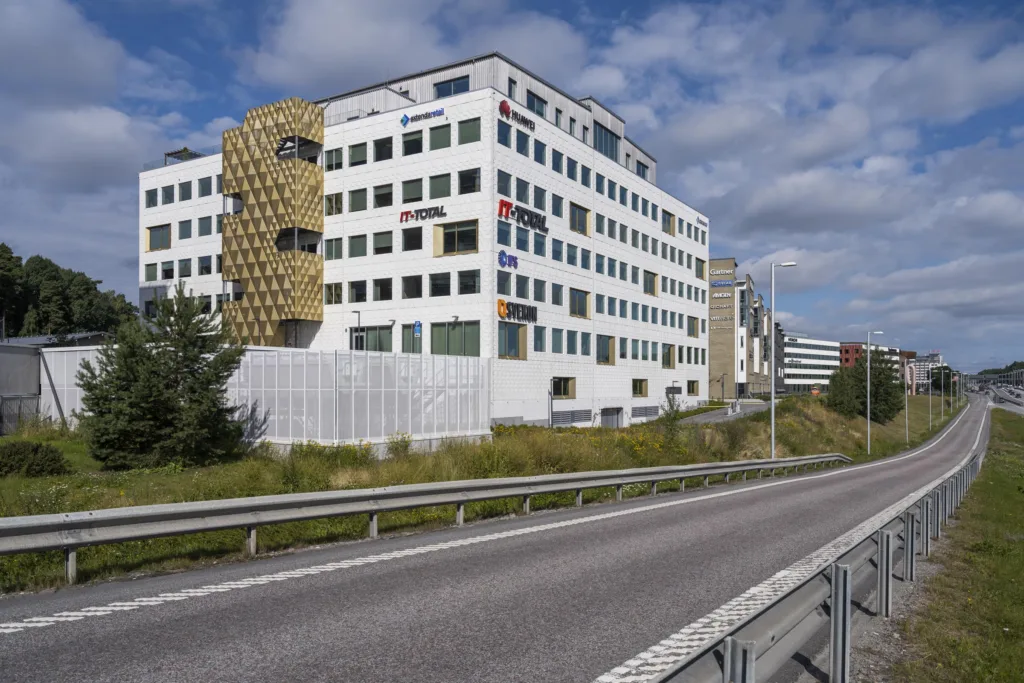 En modern vit byggnad med flera våningar, omgiven av en väg och grönska under en blå himmel.