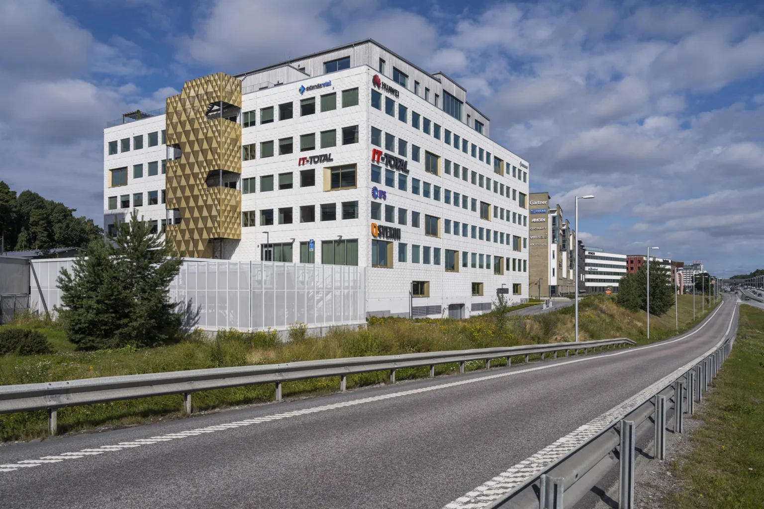 En modern vit byggnad med flera våningar, omgiven av en väg och grönska under en blå himmel.