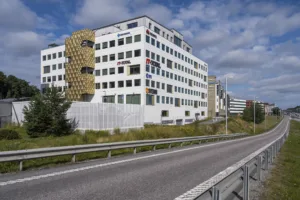 En modern vit byggnad med flera våningar, omgiven av en väg och grönska under en blå himmel.