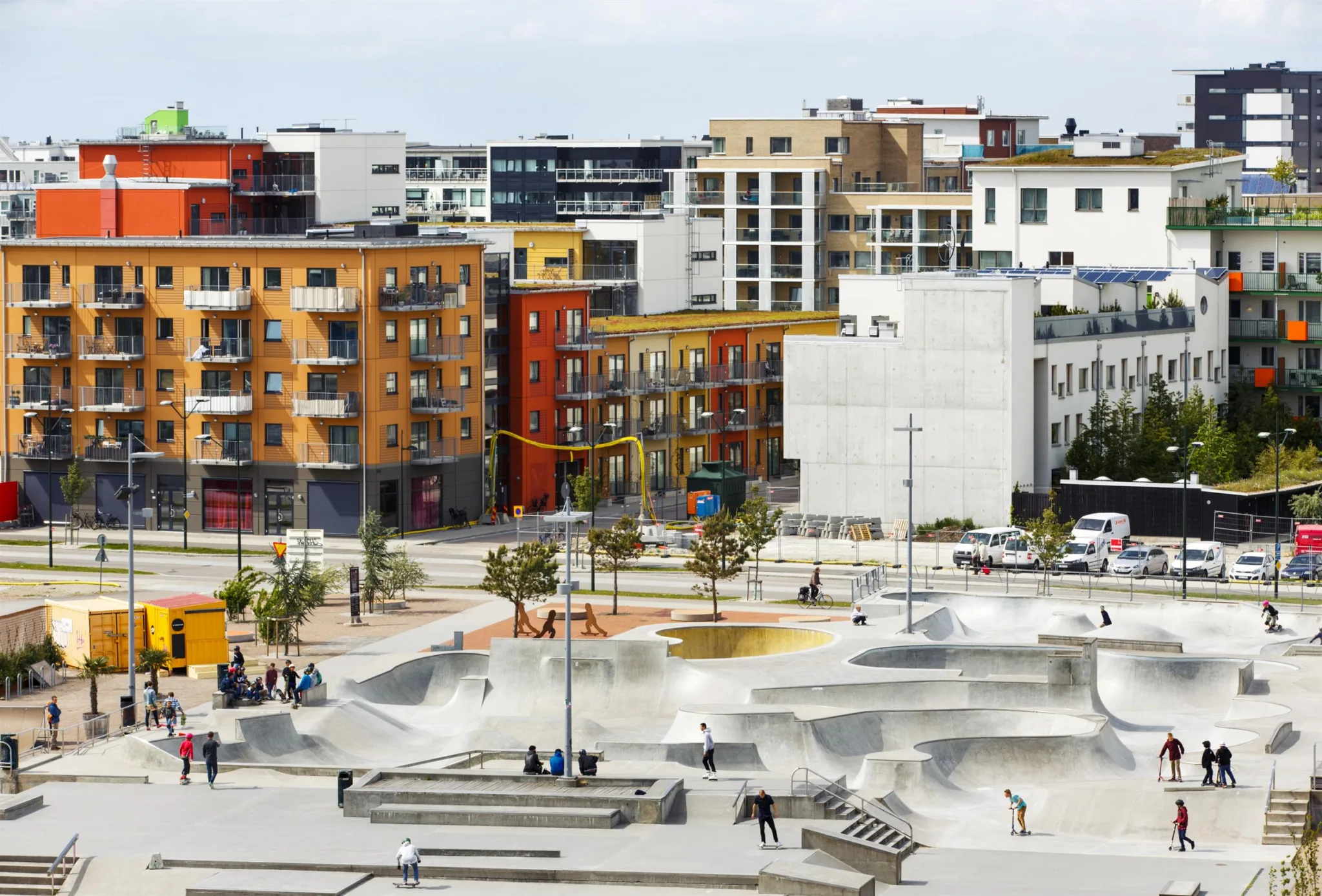 Utomhus skatepark omgiven av färgglada byggnader och träd, med människor som åker skateboard.