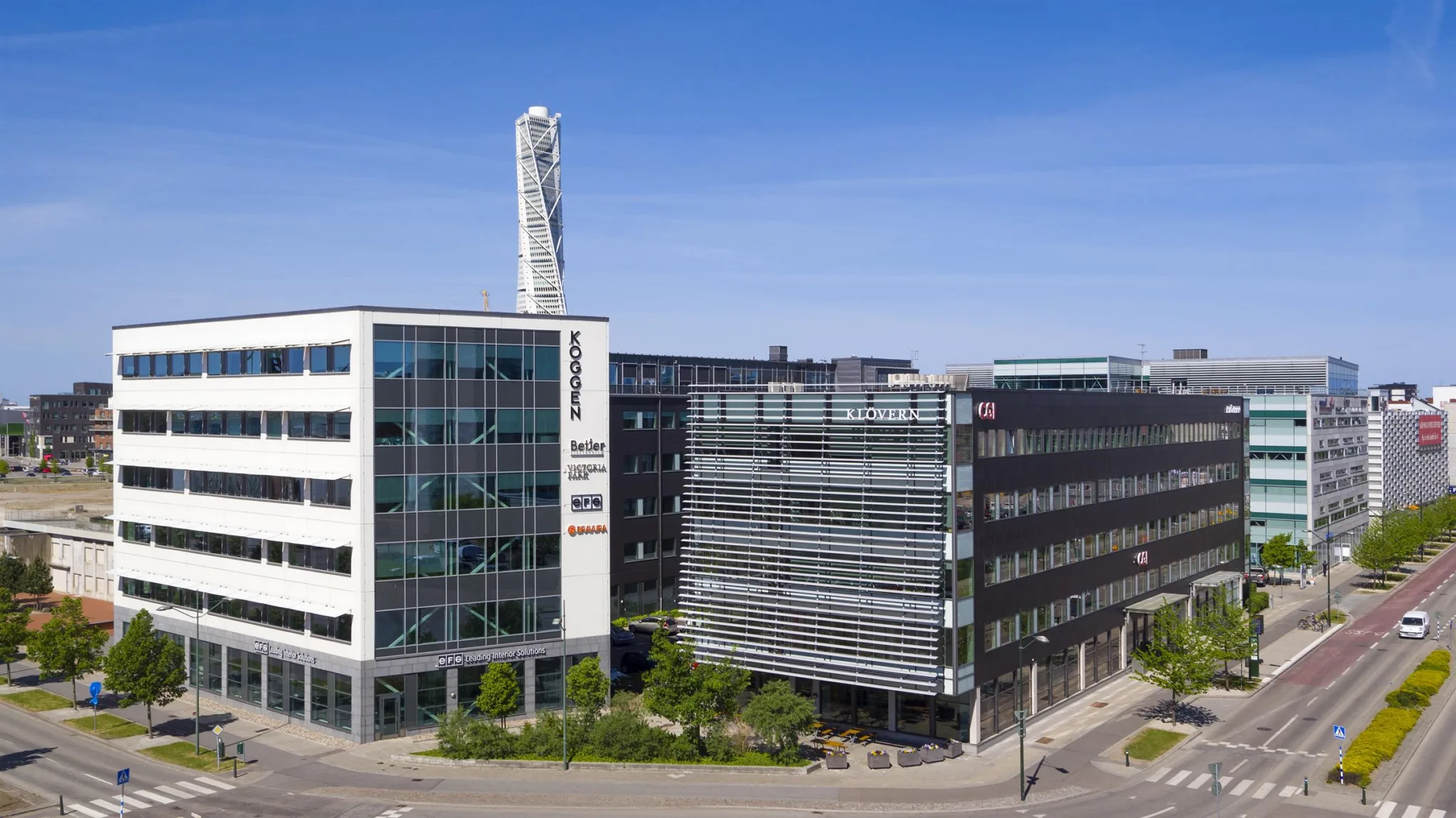 Flera kontorsbyggnader med glasfasader vid en gata, med ett högt torn i bakgrunden.