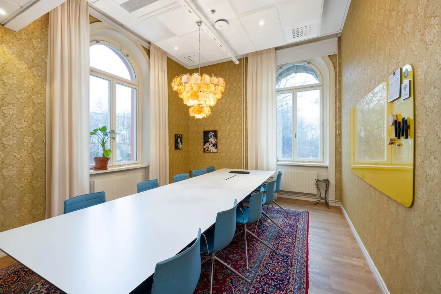 Ett konferensrum med ett långt bord, blå stolar, en ljuskrona och stora fönster med dagsljus.