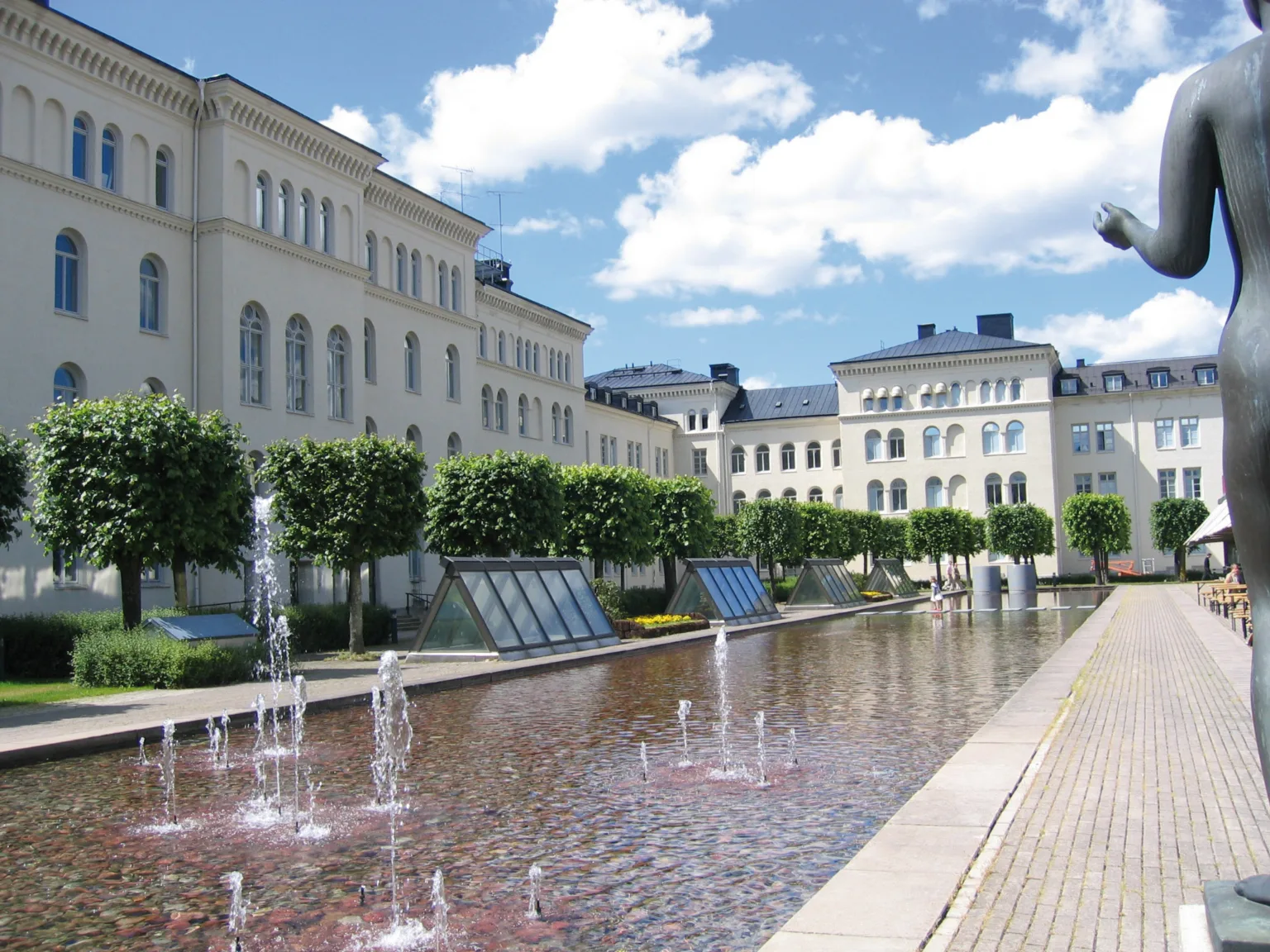 En utomhusmiljö med en fontän, träd och en rad ljusa byggnader under en blå himmel.