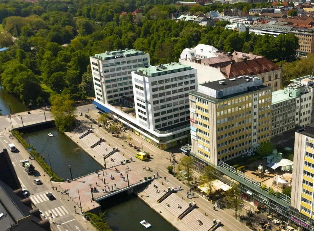 Flygvy över ett område med flera större byggnader, omgivna av grönska och en kanal med gångbroar.