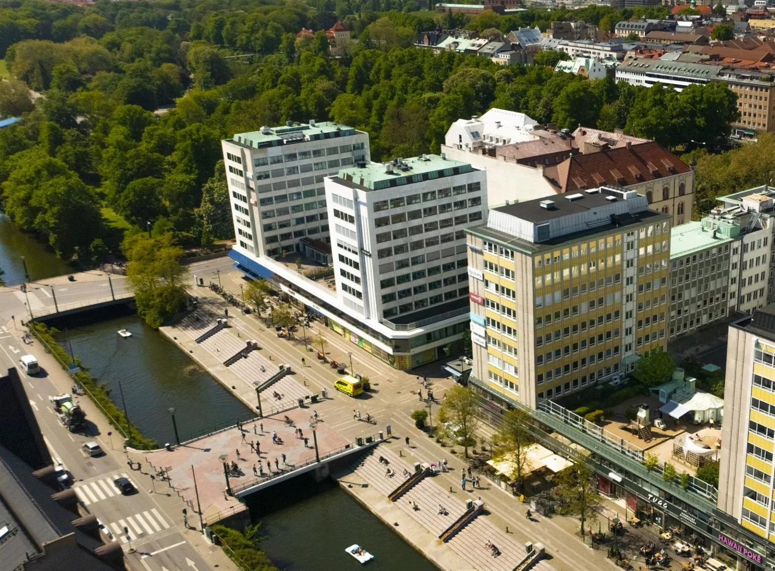 Flygvy över ett område med flera större byggnader, omgivna av grönska och en kanal med gångbroar.
