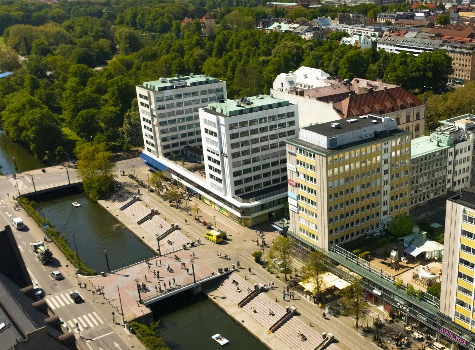 Flygvy över ett område med flera större byggnader, omgivna av grönska och en kanal med gångbroar.