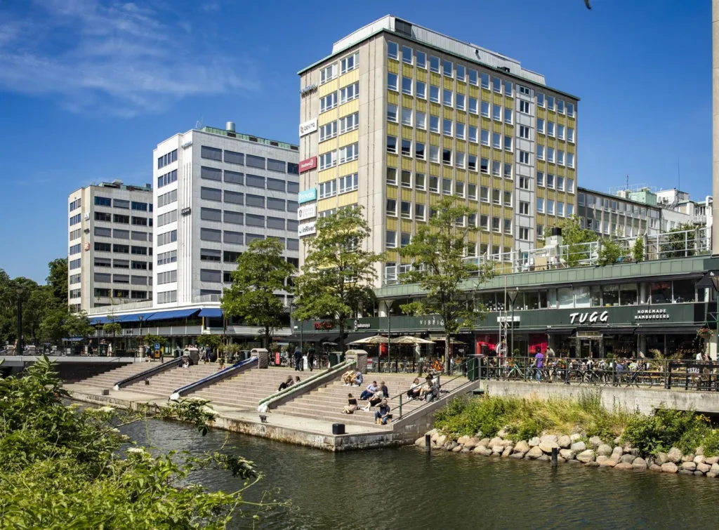 En stadsmiljö med byggnader intill en kanal, människor sitter vid trappor längs vattnet.