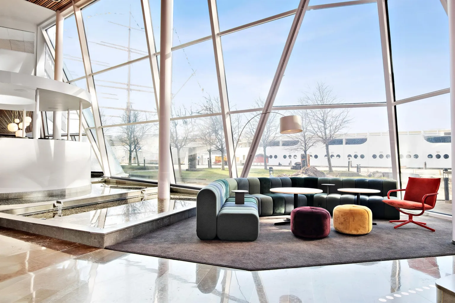 3. Loungeområde med soffor och färgglada sittpuffar vid stora fönster med utsikt över en byggnad och himmel.