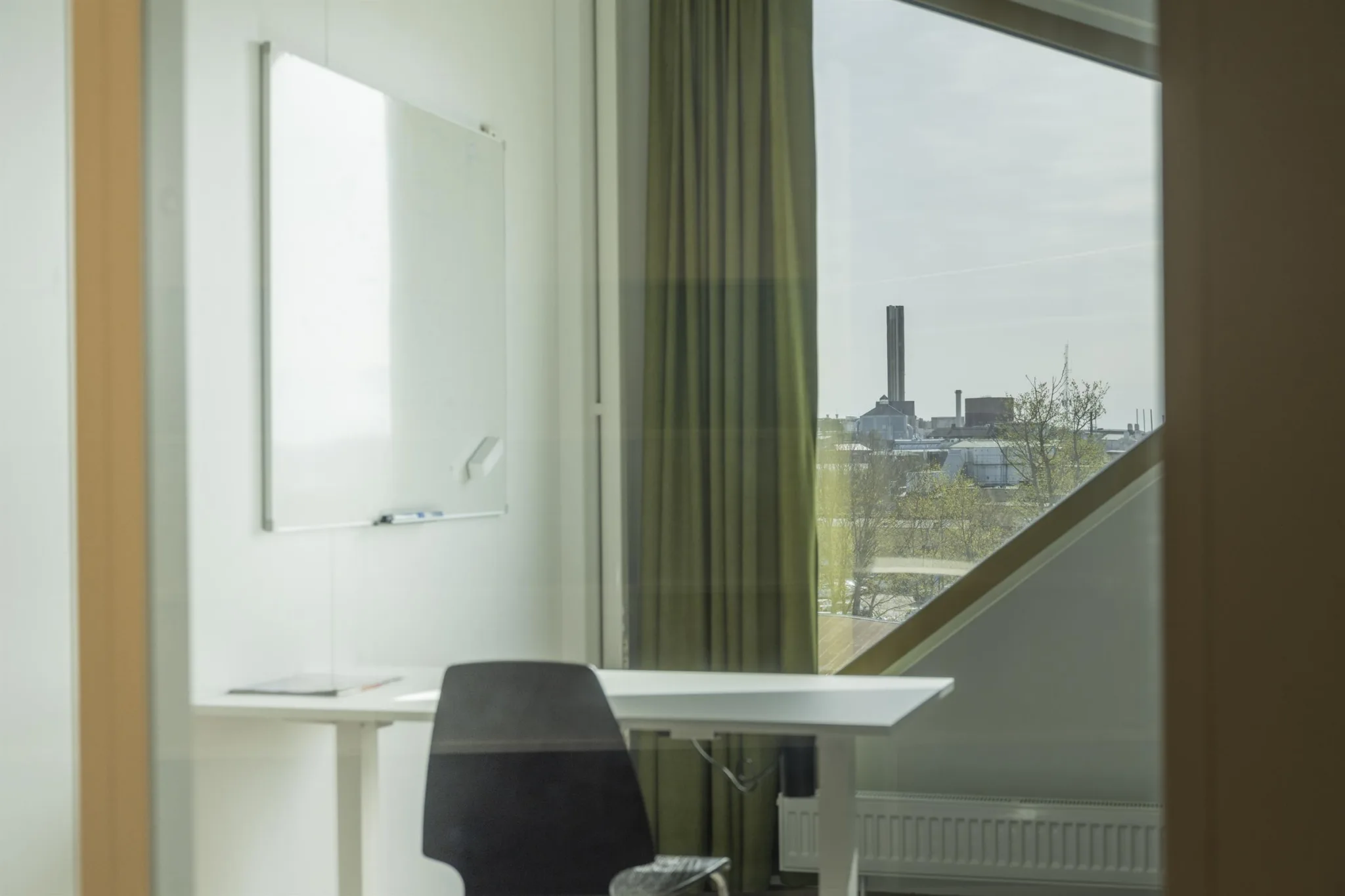 En vit arbetsplats med ett skrivbord och stol nära ett triangelformat fönster med utsikt över en fabriksbyggnad och grönska.