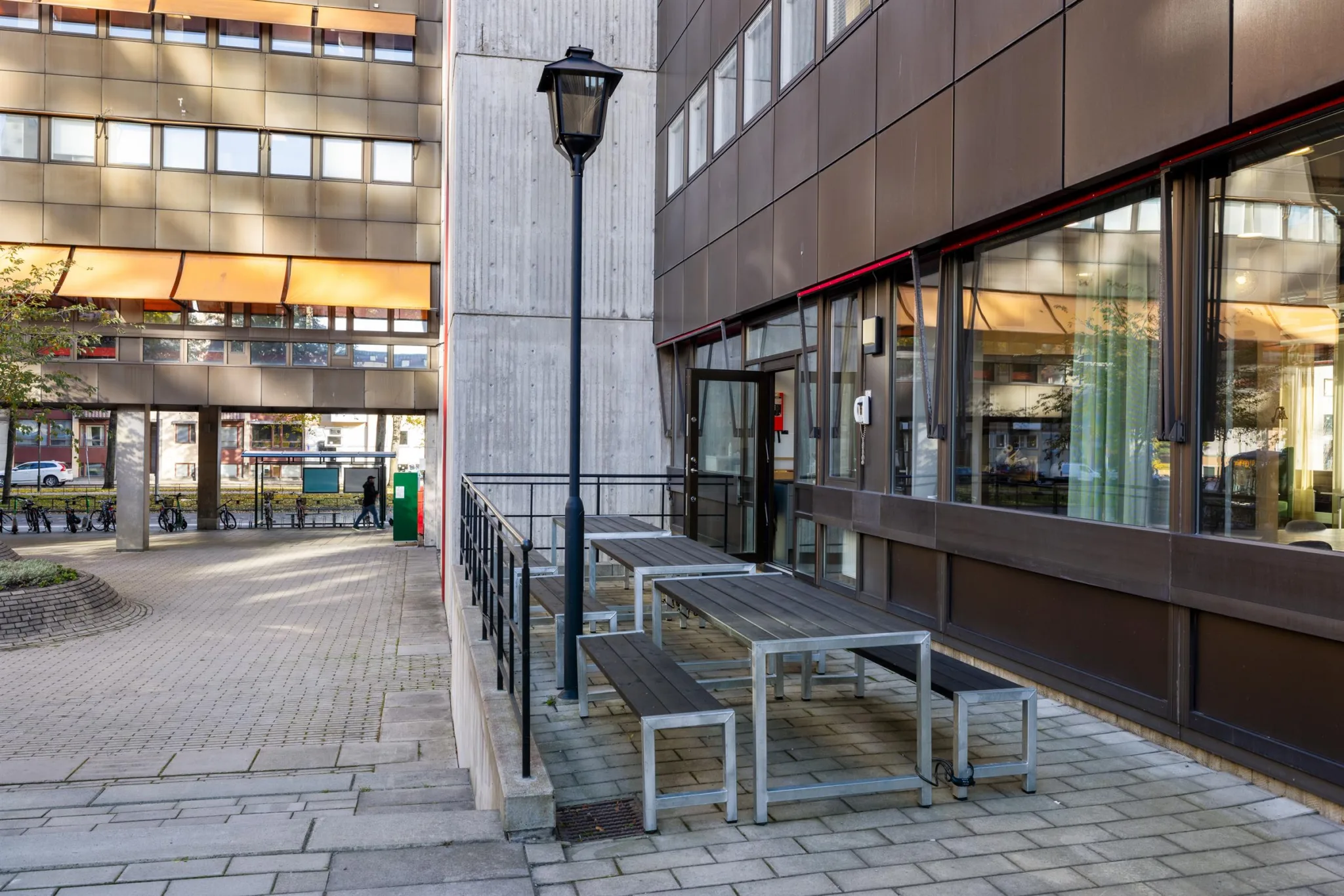 En utomhusplats med bord och bänkar i metall vid en byggnad, omgiven av trottoar och en lyktstolpe.
