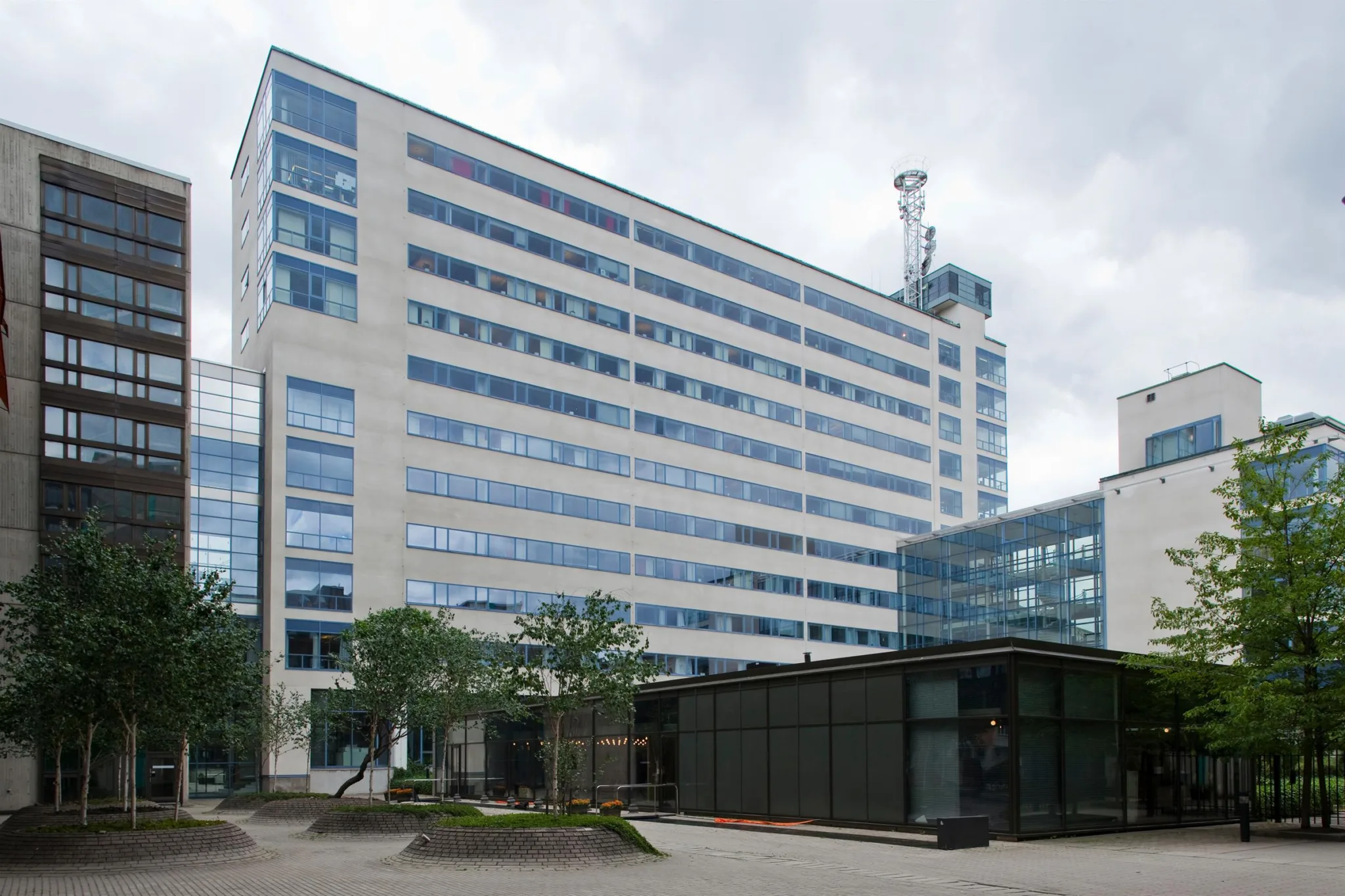 En stor kontorsbyggnad med glasfönster och flera våningar, omgiven av träd och ett mindre svart byggnad på markplan.