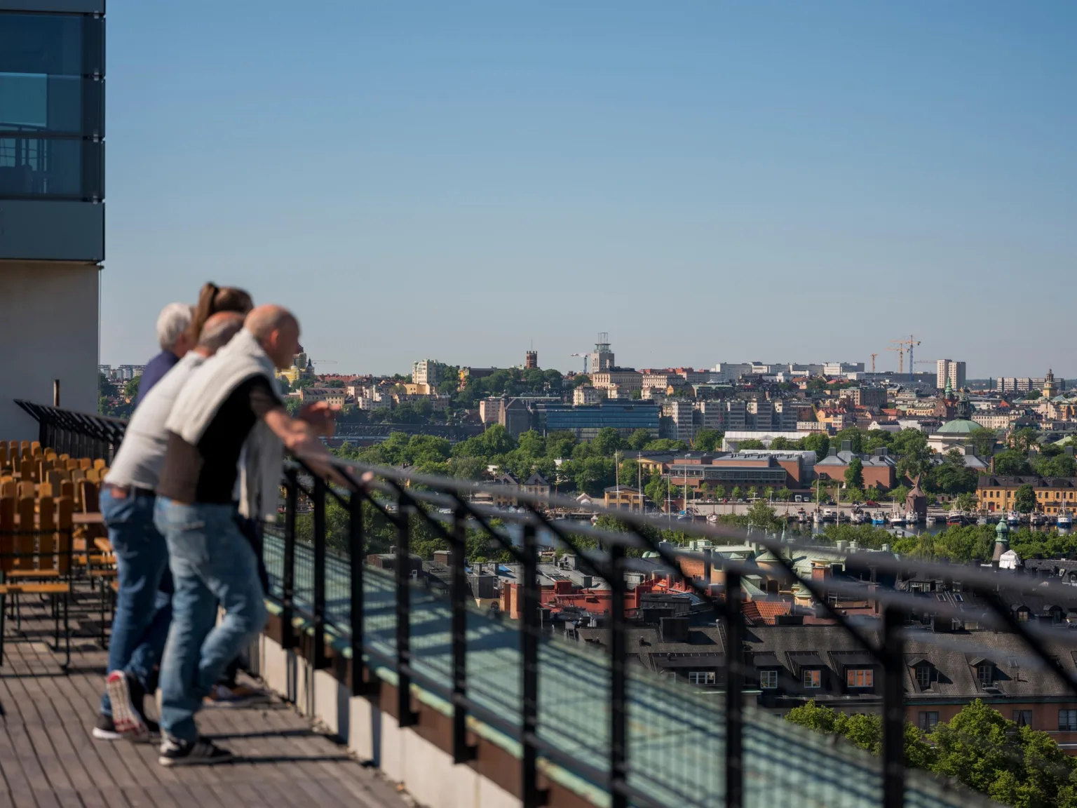 Personer står vid ett räcke och tittar ut över en stad med byggnader och grönska under en klar blå himmel.